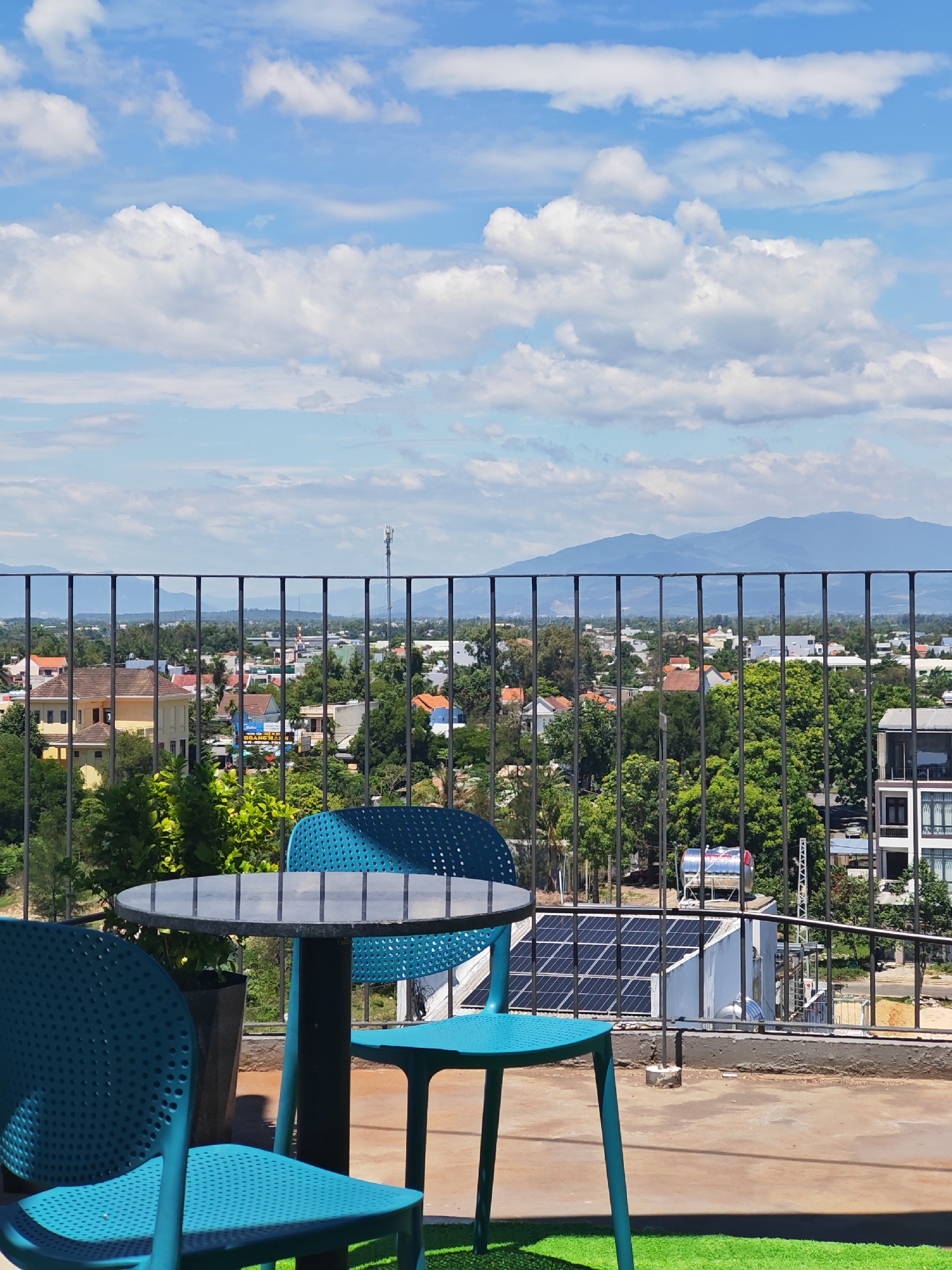 This is a full-frame shot of an outdoor seating area overlooking a cityscape in Da Nang, Vietnam. In the foreground, two turquoise chairs and a circular black table are arranged on a concrete patio with a patch of artificial turf. A black metal railing separates the patio from the view beyond. The background reveals a dense urban landscape with buildings of varying sizes and colors, interspersed with lush green trees. In the distance, a range of blue mountains stretches across the horizon under a bright blue sky dotted with white, fluffy clouds. The scene is brightly lit by natural sunlight, suggesting it is daytime. No people are visible in the image.
