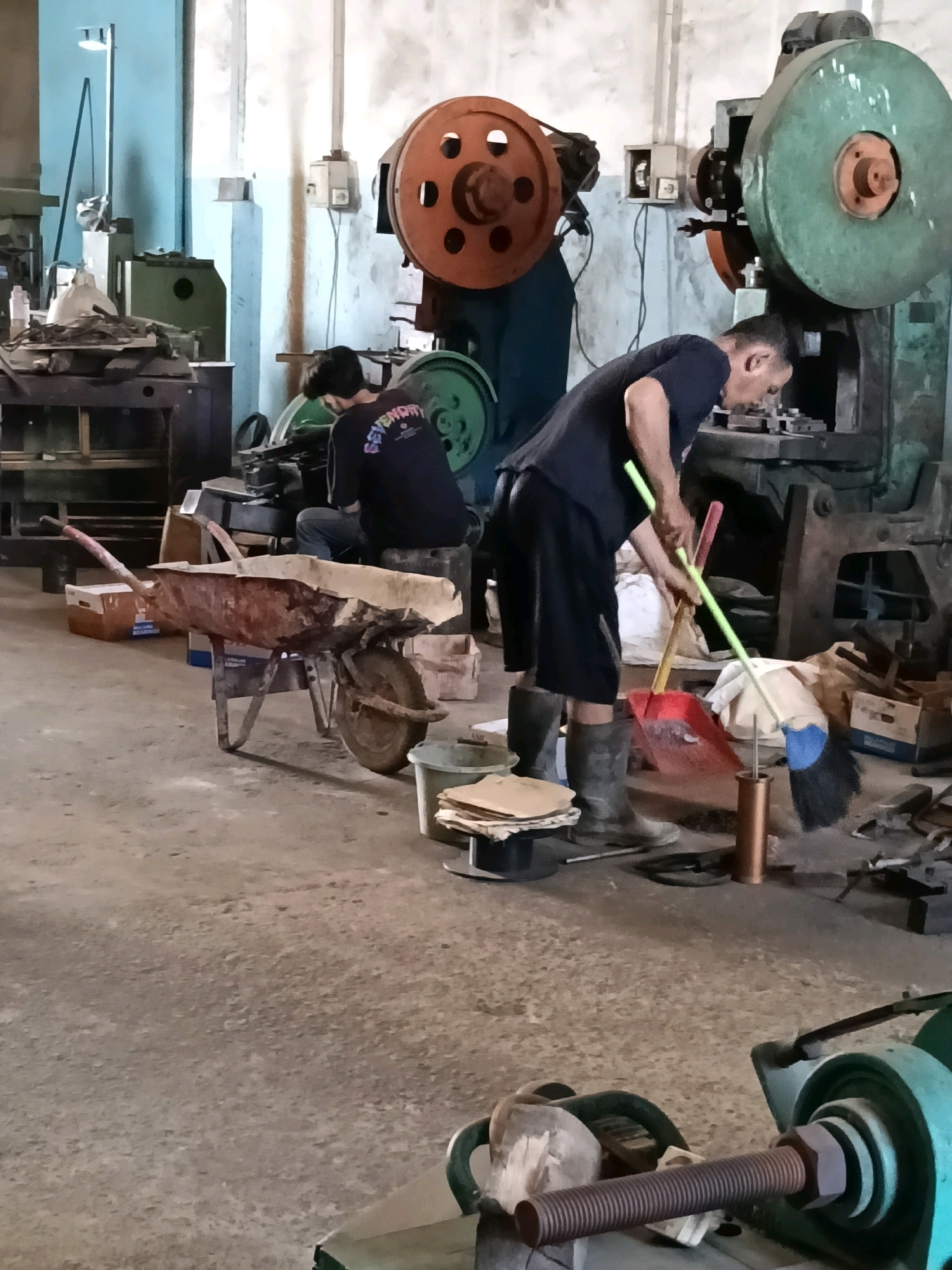 This image depicts a workshop scene in Belimbing, Indonesia, likely a metalworking or fabrication facility. The environment appears industrial with concrete flooring and exposed walls.
In the foreground, a man is actively sweeping the floor with a red dustpan and a broom. He is wearing a dark t-shirt, black shorts, and grey rubber boots. Behind him, another person is seated, appearing to be engaged in some task at a machine, wearing a dark t-shirt with lettering and jeans.
Dominating the background are large industrial machines, including what appear to be presses with prominent circular flywheels, one orange and one green. Various tools and materials are scattered around the workshop, including a rusty wheelbarrow filled with what looks like wood or scrap material, cardboard boxes, buckets, and other unidentifiable objects. The lighting suggests an indoor setting, possibly with natural light supplemented by artificial sources. The overall impression is one of an active, albeit somewhat cluttered, working environment.