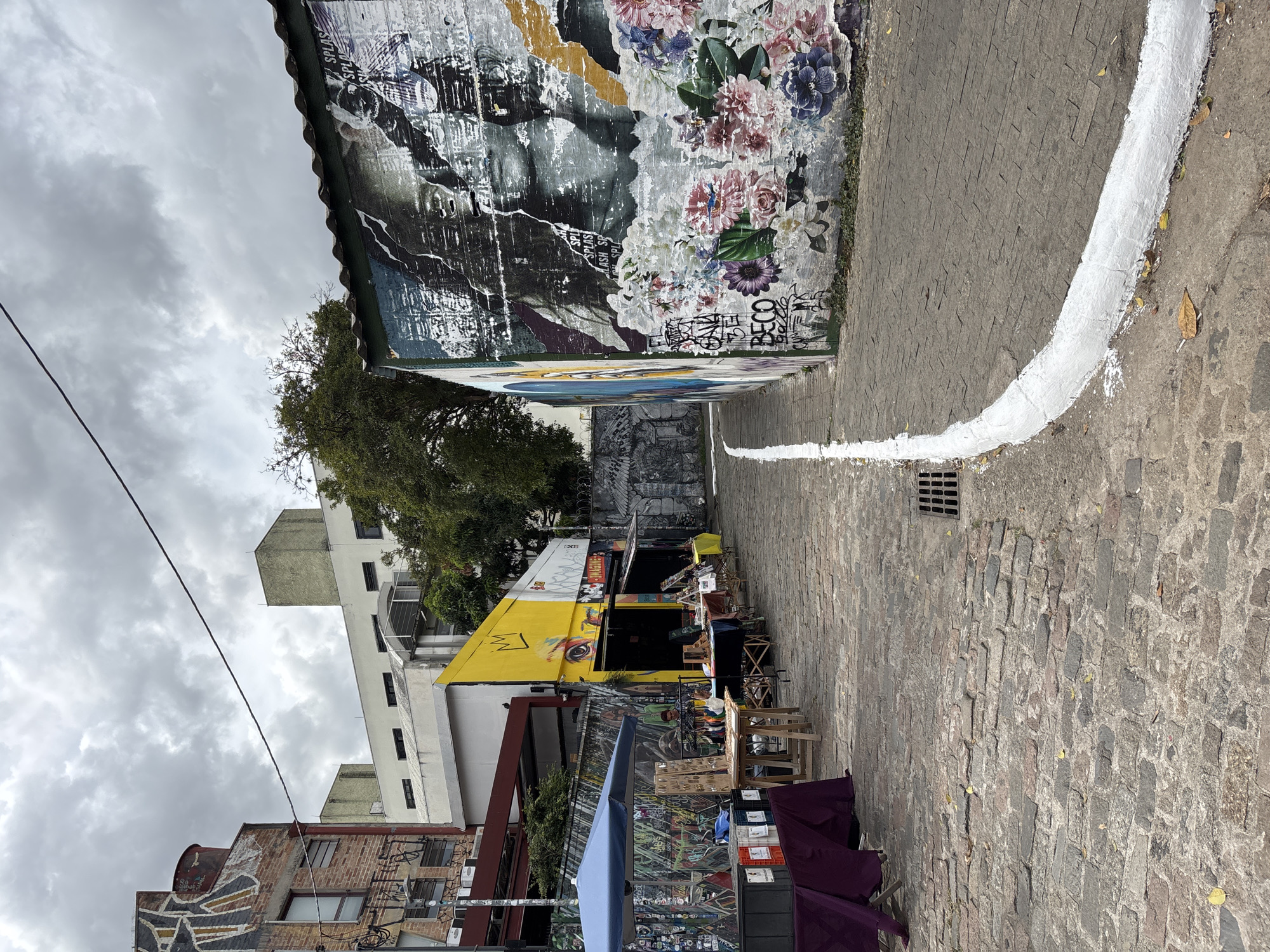 This image shows a narrow cobblestone street in what appears to be an artistic district of São Paulo, Brazil. Tall buildings line the street. The wall to the right is covered in graffiti and murals. A thick white line is painted along the right edge of the street. On the left side of the street is a line of vendors selling merchandise under tents and awnings. There is a yellow building with art on it behind the vendors' booths. The sky is overcast.