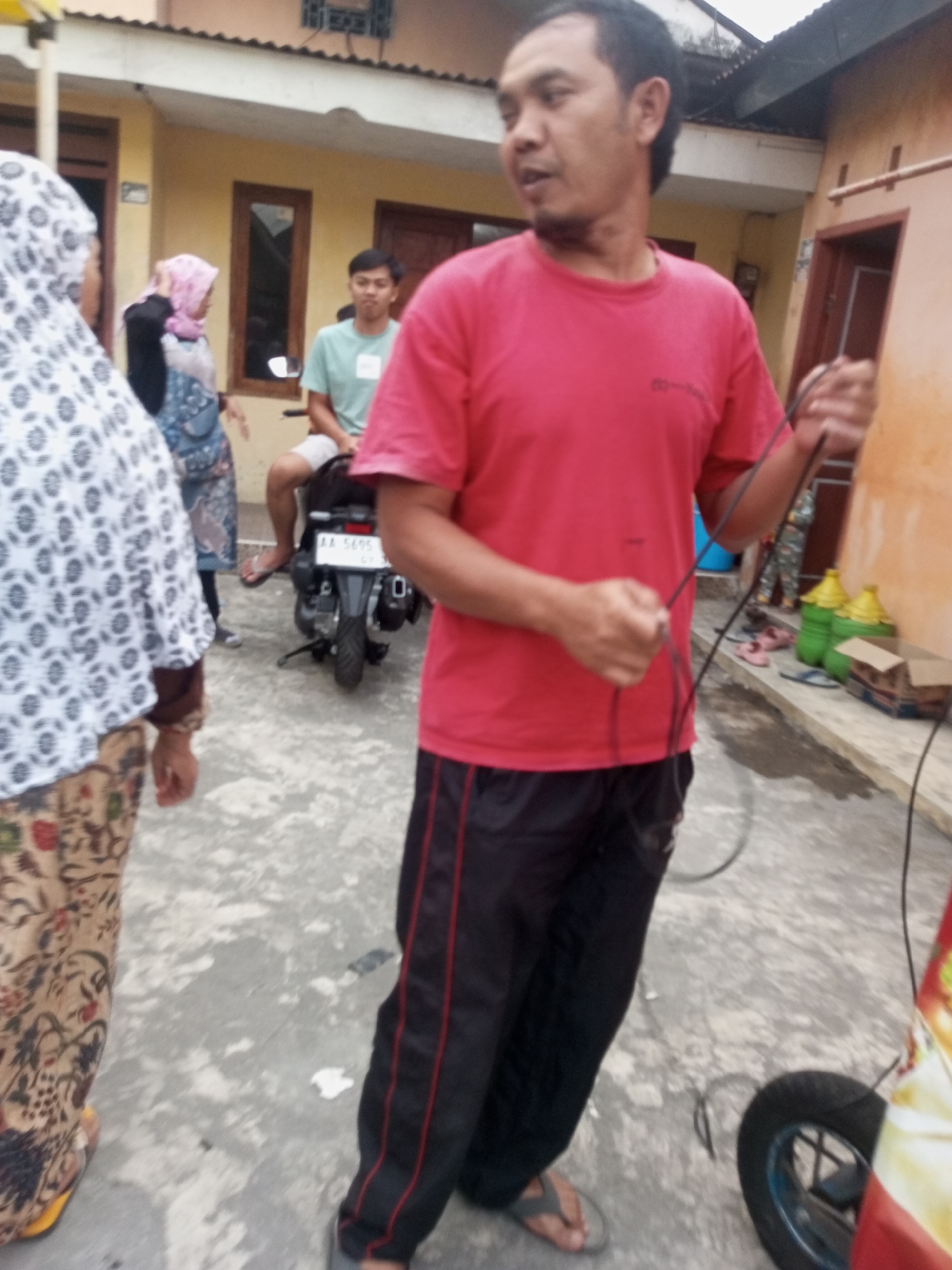 A man in a red t-shirt and black pants with red stripes stands in front of a building, holding a black cord. Behind him, a person on a motorcycle wears a light green shirt and shorts. To the left, a woman in a patterned headscarf and skirt stands facing away from the camera. In the background, two green bottles are visible. The scene appears to be an outdoor courtyard with a concrete floor.