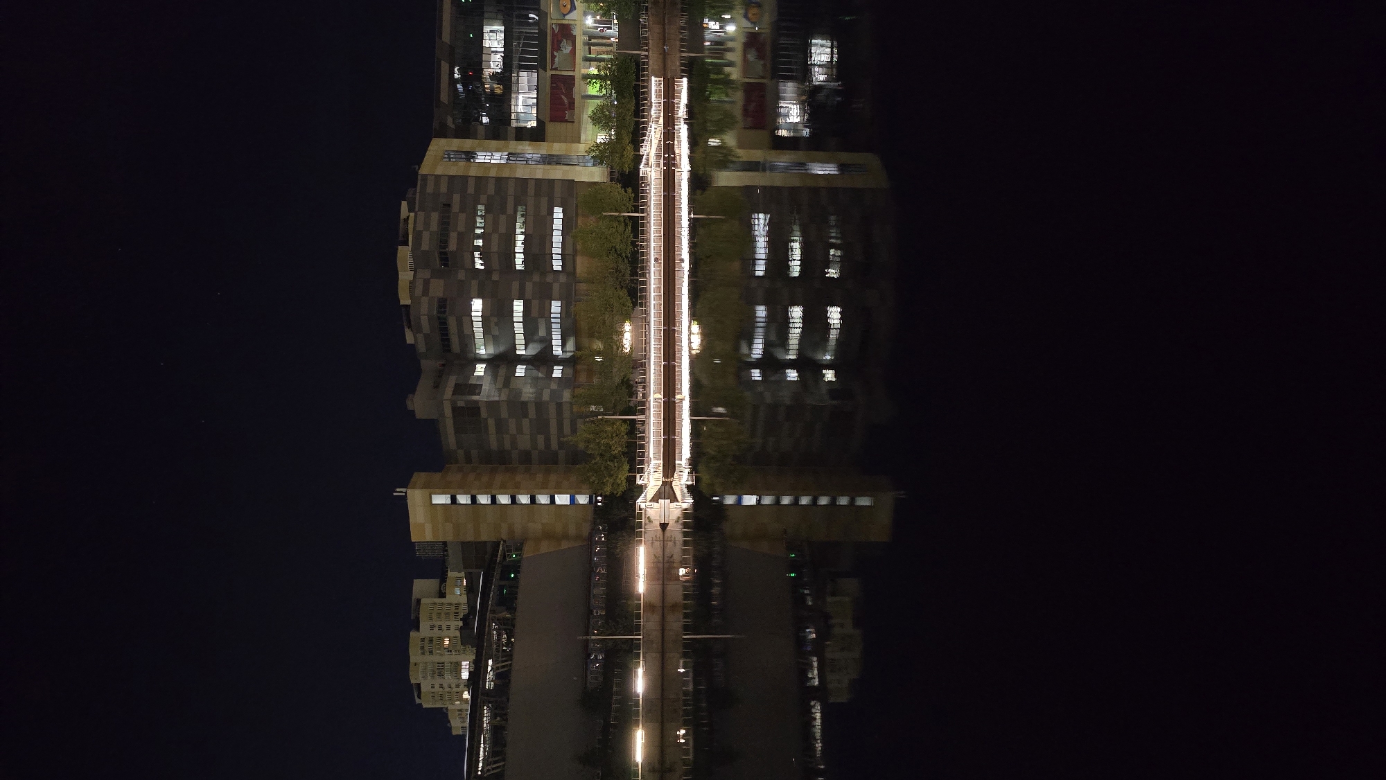 The image shows a waterfront view of modern buildings at night, with their lights reflecting in the dark water below. The buildings have a contemporary architectural style, with geometric patterns and numerous windows. A walkway with lights runs along the waterfront, separating the buildings from the water. There are trees lining the walkway. The sky above is dark, with a few faint stars visible. The overall scene depicts an urban environment at night, likely a commercial or residential district. The location is identified as Kyiv, Ukraine, implying the buildings are situated along one of the city's rivers, possibly the Dnieper. No people are visible in the image.