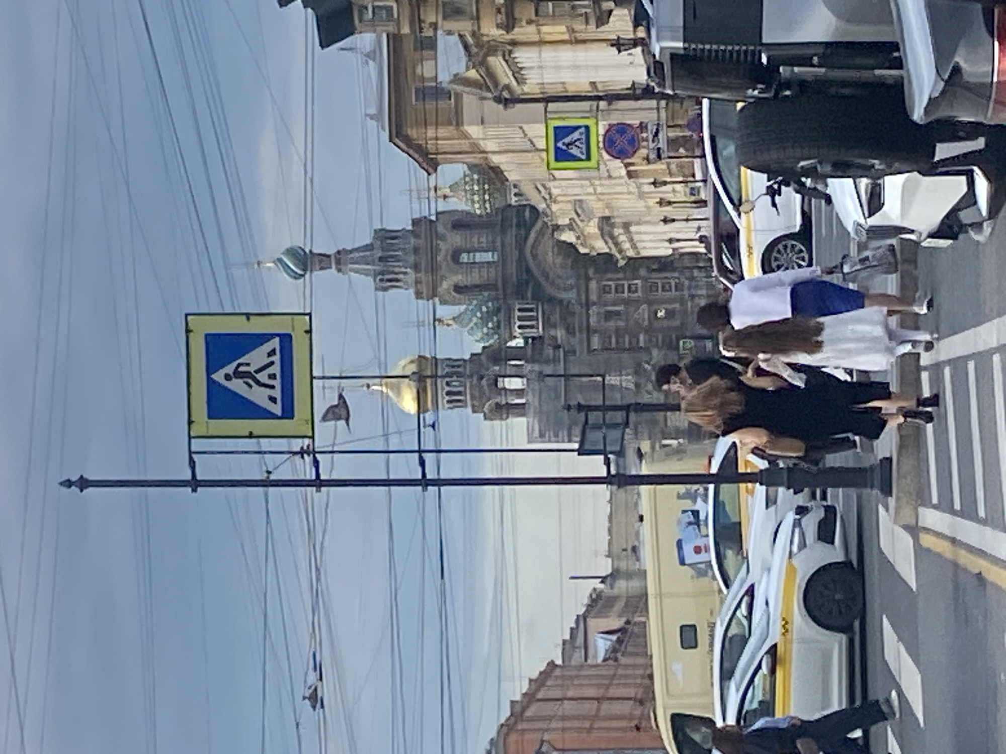 This is a photograph taken in Saint Petersburg, Russia, featuring a street scene with a prominent pedestrian crossing. In the background, the iconic Church of the Savior on Spilled Blood is visible, recognizable by its multiple colorful domes and intricate architecture. The sky is overcast, with numerous power lines crisscrossing the frame, typical of urban environments.
In the foreground, a pedestrian crossing is occupied by several individuals. To the left, a woman in dark clothing is walking away from the camera, carrying a brown bag. To her right, a young girl in a white dress is walking, with her back to the camera, followed by a woman in a white top and blue skirt. They are crossing the street alongside other pedestrians and vehicles.
Several cars are present, including white taxis with yellow stripes, a light-colored van, and a dark SUV in the foreground, partially obscuring the view. Traffic signs, including a pedestrian crossing sign with a blue background and a white silhouette of a person walking, are mounted on a pole. The street is paved, and markings for the pedestrian crossing are clearly visible. The overall atmosphere suggests a busy urban street in Saint Petersburg, likely during daylight hours.