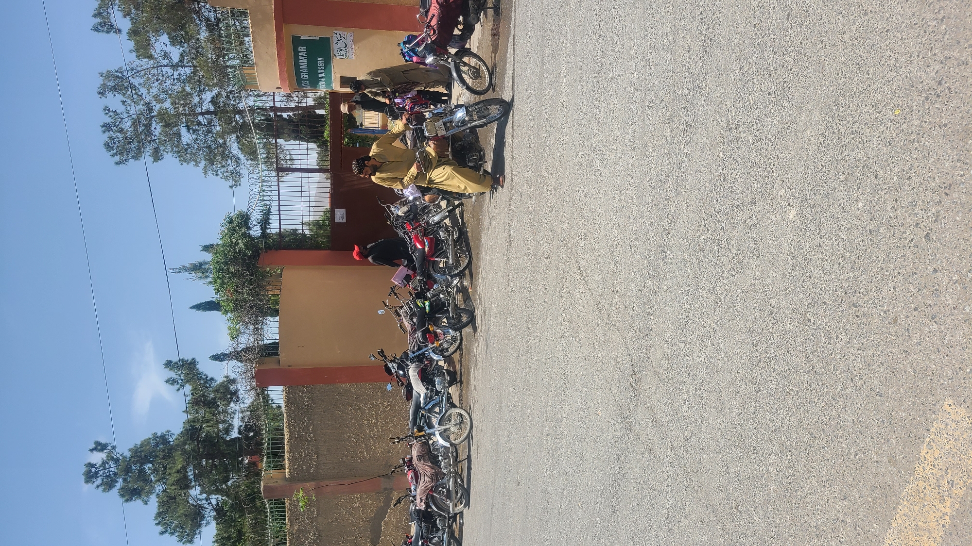 The image depicts a row of motorcycles parked along a street in Quetta City Tehsil, Pakistan. The bikes are parked next to a building with a sign that reads "Grammar Nursery." Several people are present near the motorcycles, some sitting on them. The street is paved and appears to be in daylight. The sky is blue and clear. Trees are visible in the background.