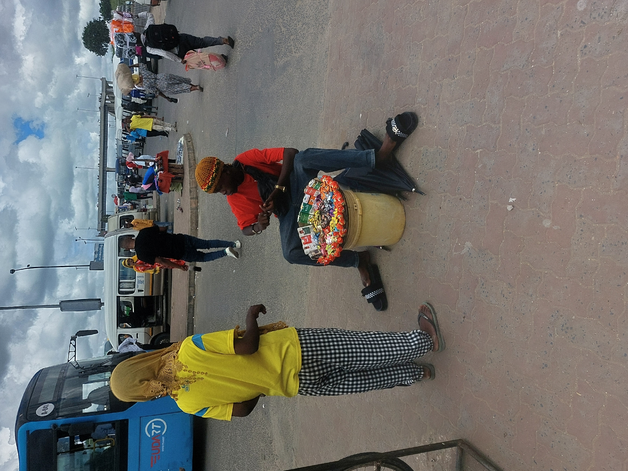 The image depicts a street scene in Dar es-Salaam, Tanzania. A man wearing a yellow and orange beanie, a red t-shirt, and blue jeans is sitting on the ground, with a bucket filled with candy and cigarettes in front of him. An open umbrella is visible next to him. A woman in a yellow top, a beige headscarf, and black and white checkered pants stands near the man. A blue bus with "UDART" written on the side is parked nearby. Other people are walking around in the background, and a row of white vans with open doors lines the street. There is also an elevated walkway over the road and an overcast sky above.
