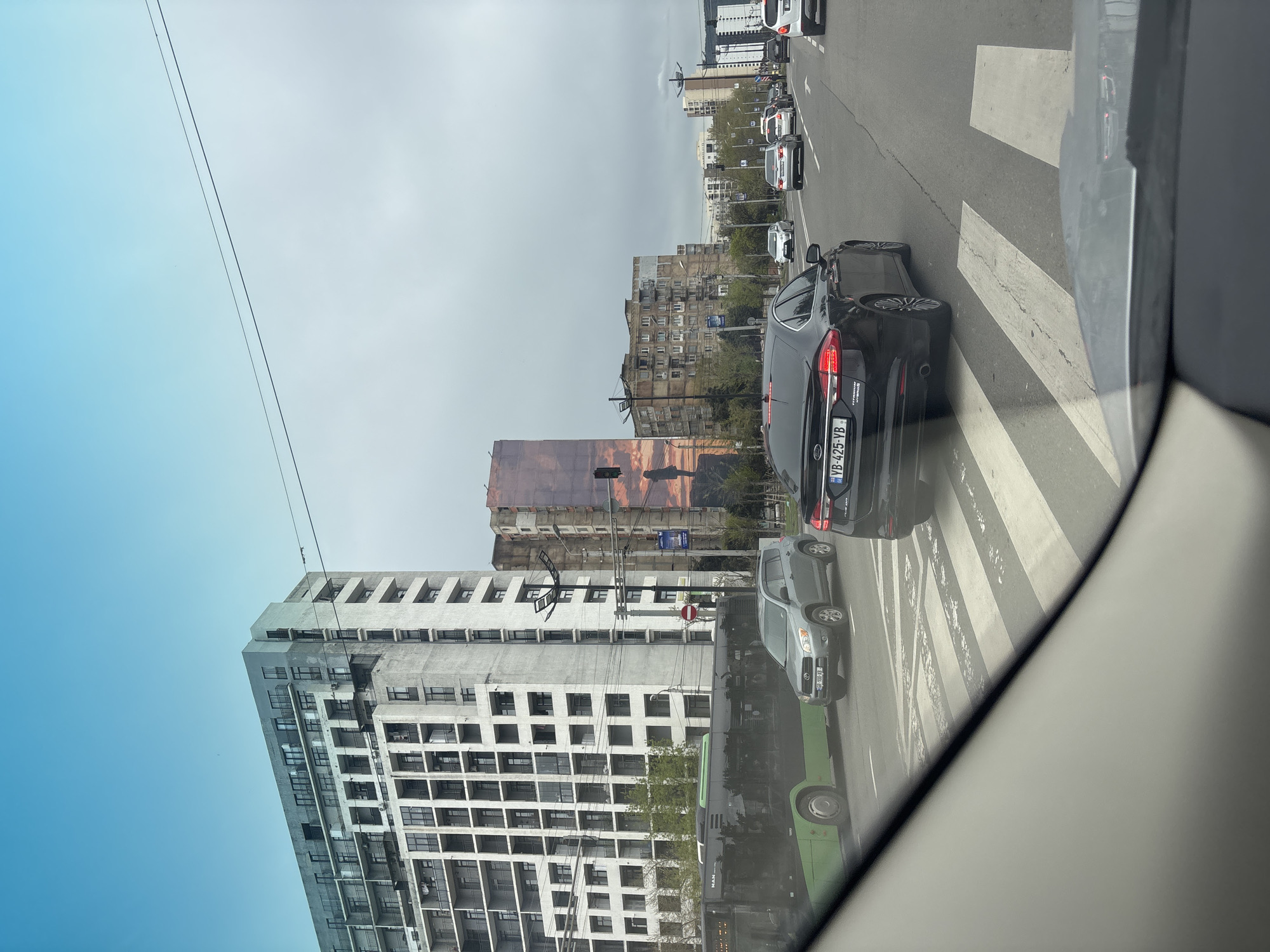 The image captures a street scene in Tbilisi, Georgia, from the perspective of inside a vehicle, likely looking out the front passenger side. The sky is partly cloudy, with a clear blue patch on the left gradually transitioning to a light grey overcast on the right. Overhead power lines traverse the upper portion of the frame.
In the foreground, the frame of the vehicle taking the picture is visible on the right. A multi-lane road with a zebra crossing is prominent, showing wear and tear. Several cars are present, suggesting traffic. A black sedan with a Georgian license plate "YB-425-VB" is stopped near the zebra crossing, followed by a grey SUV with the license plate "KB-900-KB". Behind the SUV, a green public bus with the "MAN" logo is visible. More cars are queued further down the road.
The street is lined with buildings of varying architectural styles. On the left, a tall, modern building with a grey facade featuring many square windows and balconies dominates the lower part of the frame. Further down the street, an older, seemingly residential building with numerous smaller windows and a somewhat weathered appearance stands. One side of this older building features a large, indistinct mural or advertisement with warm, earthy tones. Other buildings are visible in the distance, contributing to an urban environment. A red circular sign with a white horizontal bar (likely a "no entry" sign) is visible on a pole near the street.