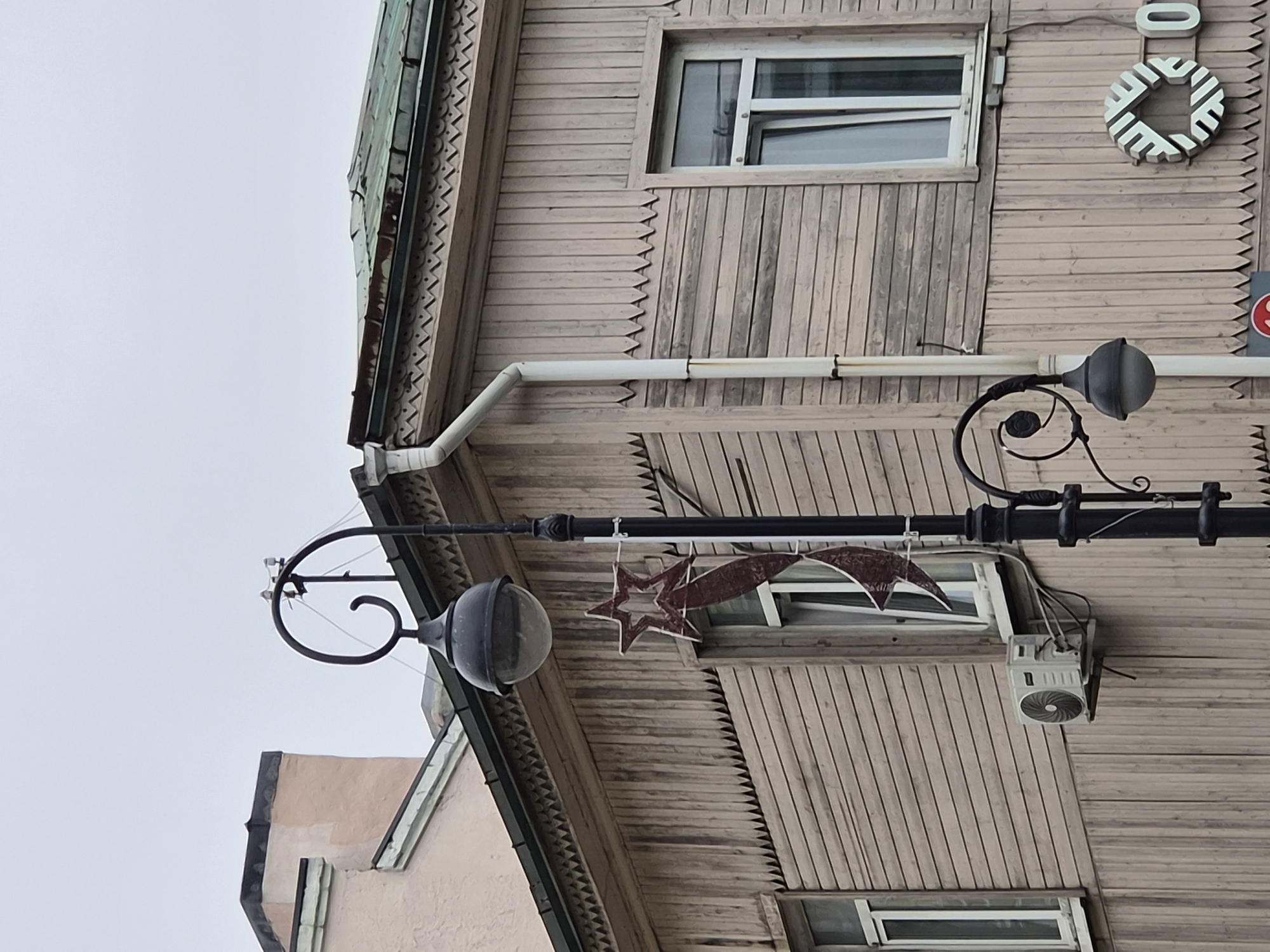 The image shows a street scene in Vladivostok, Russia, featuring a weathered wooden building with distinctive ornamental trim along the roofline. A black lamppost with a spherical glass lantern stands in front of the building, adorned with a red star decoration. Several windows are visible on the wooden structure, one of which has an air conditioning unit mounted below it. The sky is overcast.