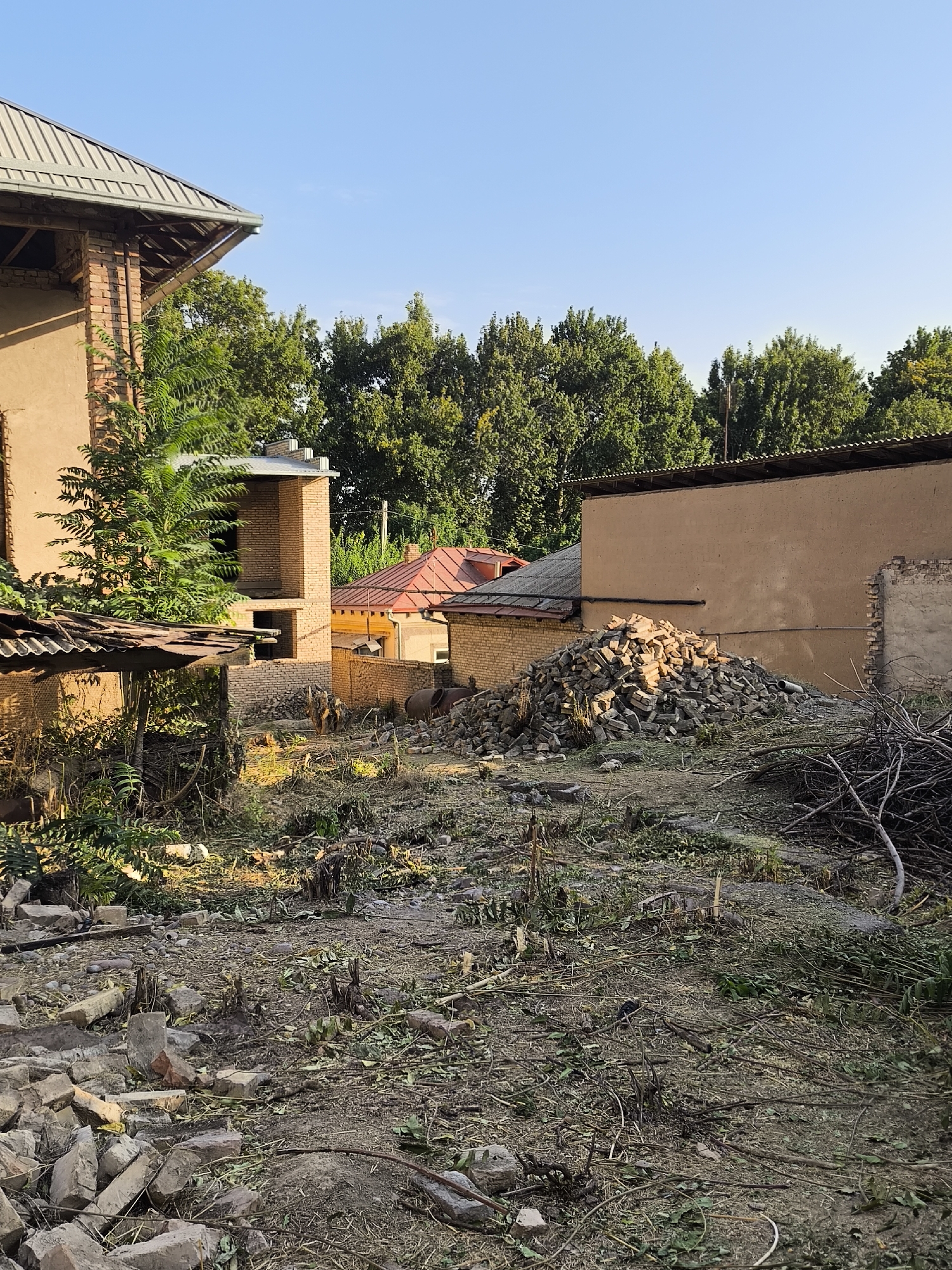 In a rural setting with clear blue skies, an outdoor scene depicts the aftermath of demolition and debris. A partially constructed brick building with a metal roof is on the left, and a larger, tan-colored building with a flat roof occupies the right side. In the foreground, a pile of broken bricks and rubble is prominent, mixed with fallen leaves, branches, and tree stumps, suggesting recent clearing or demolition work. Several smaller structures with corrugated metal roofs are also visible. The overall impression is one of disarray and ongoing construction or renovation in a yard area.