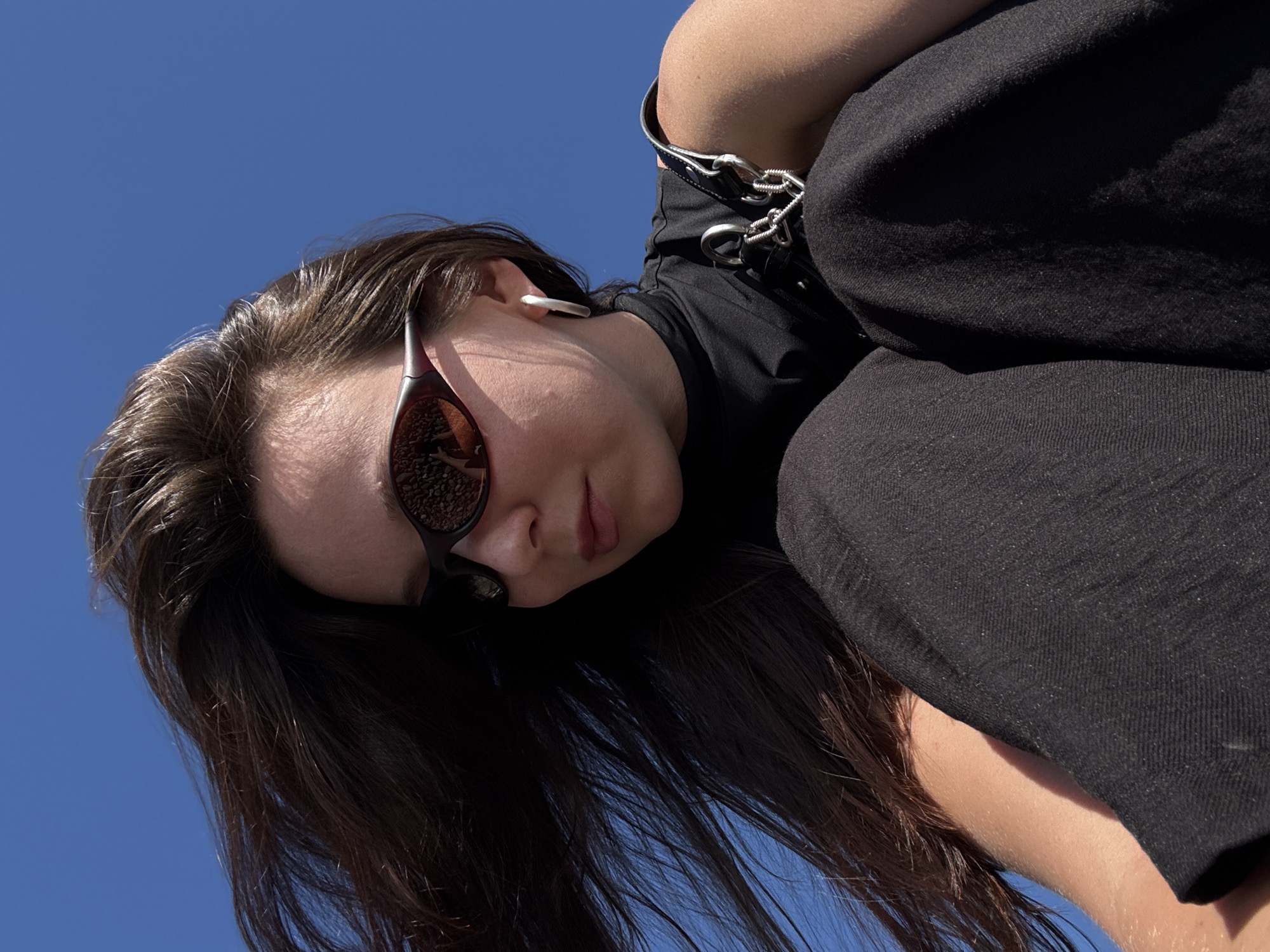 A young woman with long, dark hair and sunglasses is pictured against a clear blue sky. She is wearing a black sleeveless top with a high neckline and black pants. A silver hoop earring is visible on her left ear, and a black strap with chain detailing rests on her right shoulder. Her hair is blowing to the left, away from her face. The image appears to be a low-angle shot, possibly taken outdoors. There is no indication of the city of Sochi, Russia, in the image.