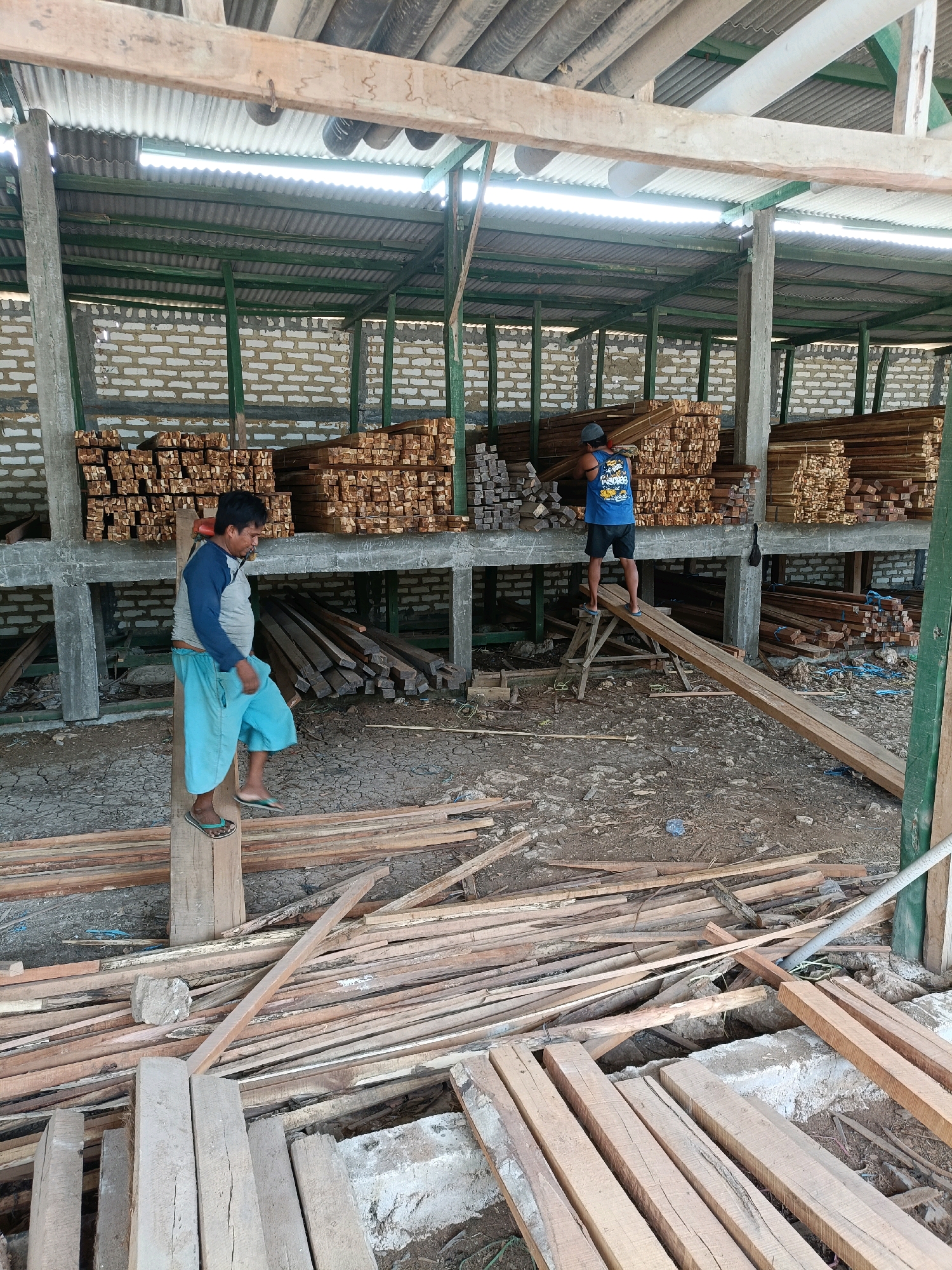 This image depicts a lumber yard in Pordapor, Indonesia. In the foreground, a man wearing a blue and gray long-sleeved shirt and light blue shorts stands on a wooden beam, with his left foot resting on a vertical wooden post. He is wearing flip-flops. To the right and in the background, another man, wearing a blue tank top with a graphic design and dark shorts, stands on a wooden plank supported by sawhorses, holding a long piece of lumber.
The environment is an open-air lumber storage area under a corrugated metal roof supported by green metal beams and concrete pillars. The walls are constructed of light-colored brick. Stacks of various sizes of lumber are visible on shelves and on the ground. The ground is covered with dirt and scattered pieces of wood. The lighting suggests it is daytime, with sunlight filtering through gaps in the roof structure.