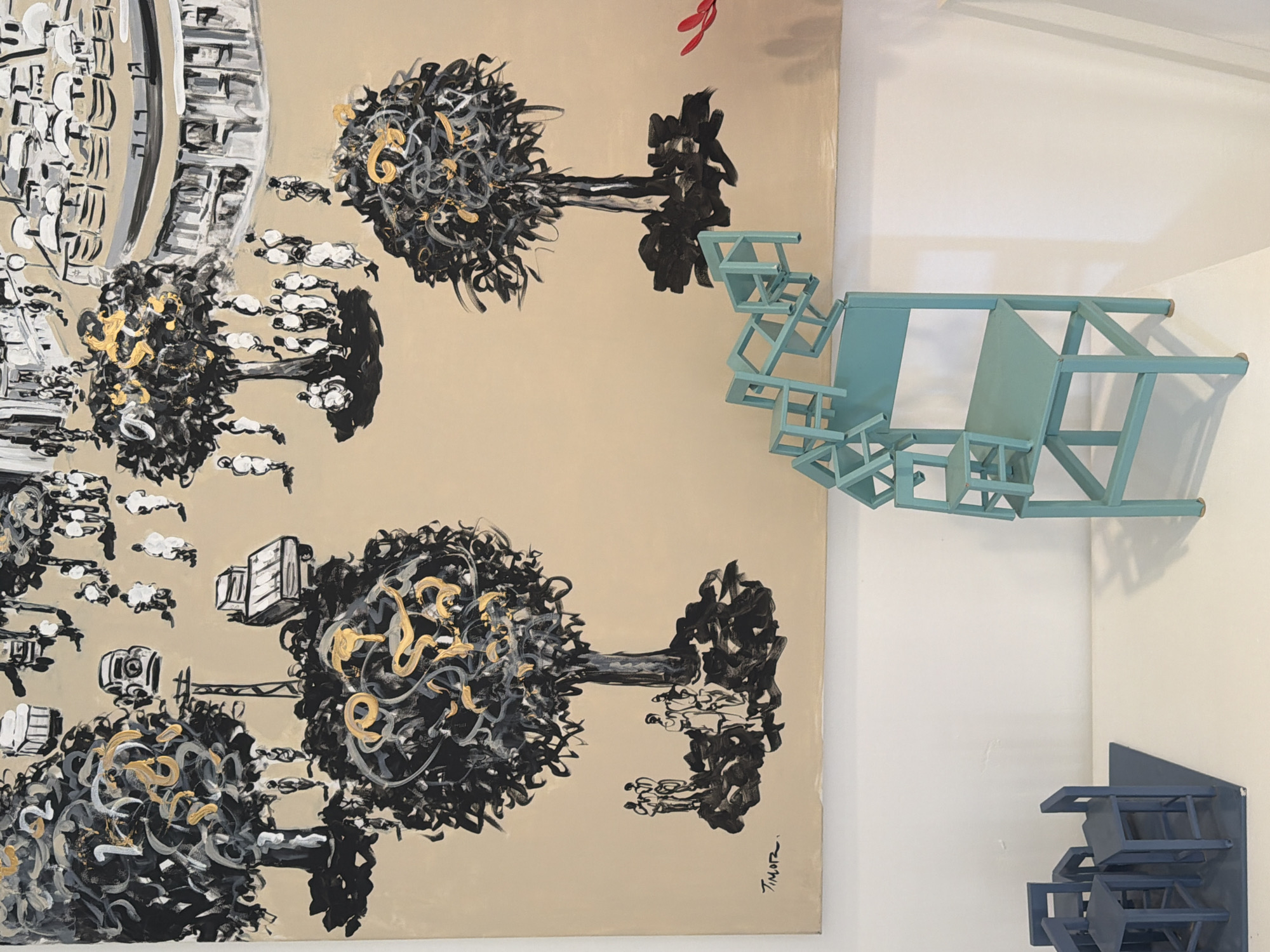 The image captures a gallery setting in Tel-Aviv, Israel. On a wall is a painting with a beige background featuring black, white, and gold abstract trees, buildings, and figures. To the right of the painting is a blue sculpture made of miniature chairs stacked and arranged in an angular pattern. Below the blue sculpture, closer to the corner of the wall, there is a smaller, dark blue sculpture composed of miniature chairs. The scene is well-lit, highlighting the textures and colors of the artwork.
