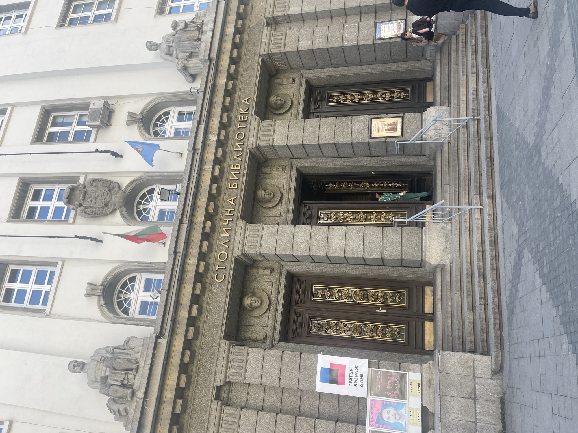 The image depicts the facade of the Столична библиотека (Sofia Library) in Sofia, Bulgaria. The architecture includes a symmetrical design with three sets of double doors leading into the building, each accessible by stairs. The building's facade is decorated with stone sculptures and reliefs. Text on the building is "СТОЛИЧНА БИБЛИОТЕКА," the library's name. There are several posters near the lower entrance. People are on and near the steps leading to the library's entrances.