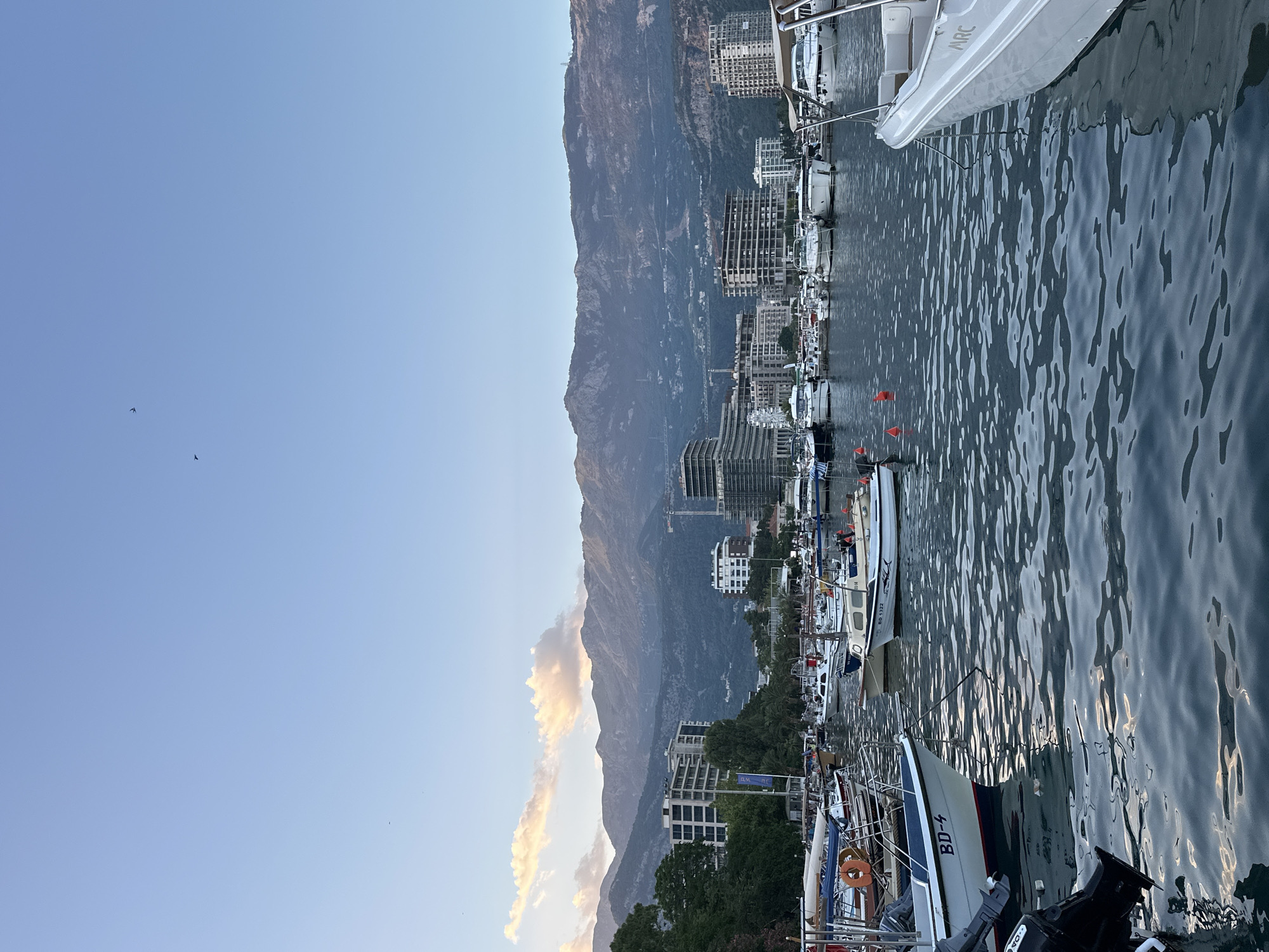 This is a daytime, eye-level, medium shot of a marina in Budva, Montenegro. The sky is a clear, pale blue with a few wispy clouds near the horizon. In the distance, a range of large, rugged mountains forms the backdrop. In the middle ground, a city skyline with modern buildings, some under construction, stretches along the coast. A ferris wheel is visible amongst the buildings. The foreground is dominated by the water of the marina, with numerous boats of various sizes moored. The water is choppy, reflecting the sky and surrounding objects. Several small to medium-sized motorboats are clearly visible, some with their outboard motors attached. One boat in the foreground has the registration "BD-4" visible. Another boat has "BD 1322" visible. There are no people visible in the image.