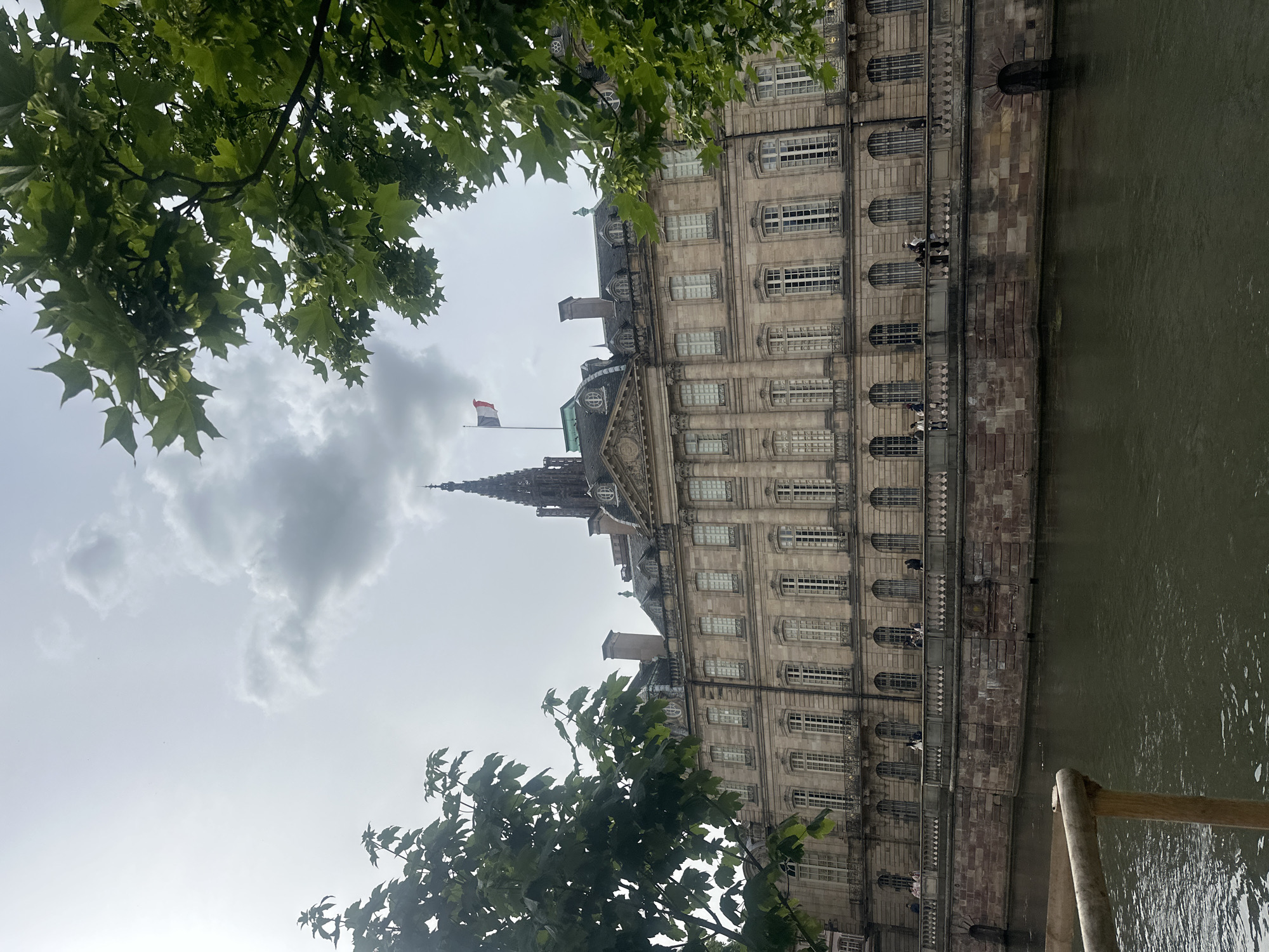 The image depicts a wide shot of the Palais Rohan in Strasbourg, France, situated along the Ill River. The grand stone building, characterized by its symmetrical architecture, numerous windows, and ornate detailing, dominates the mid-ground. A French flag flies from a flagpole on the roof. In the foreground, the dark, rippling water of the river occupies the bottom portion of the frame, with a wooden railing visible on the left. The riverbank is constructed of stone. Several individuals are visible strolling along the terrace in front of the Palais. Lush green foliage from trees partially frames the scene from the top and sides. The sky is overcast with a mix of clouds and brighter patches.