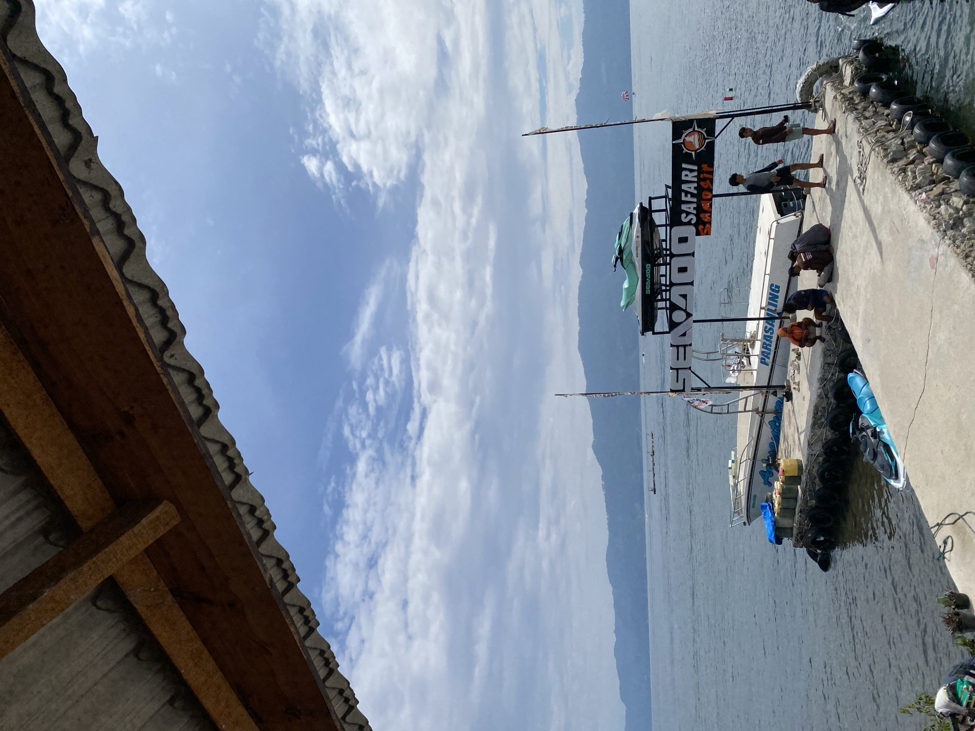 The image depicts a waterfront scene at Tuk Tuk, Indonesia. A concrete pier extends into a calm body of water, with several people present on it. On the pier, a group of individuals are gathered near a boat that has "PARASAILING" written on its side. Another group of people are standing further down the pier. A sign with "SENODO SAFARI SAMOSIR" is visible.
Two jet skis are present; one is parked on the pier and another is in the water. In the background, a range of verdant, sloping hills forms the horizon, with a hazy sky overhead dotted with clouds. A portion of a corrugated roof structure is visible in the foreground, casting a shadow over part of the scene.