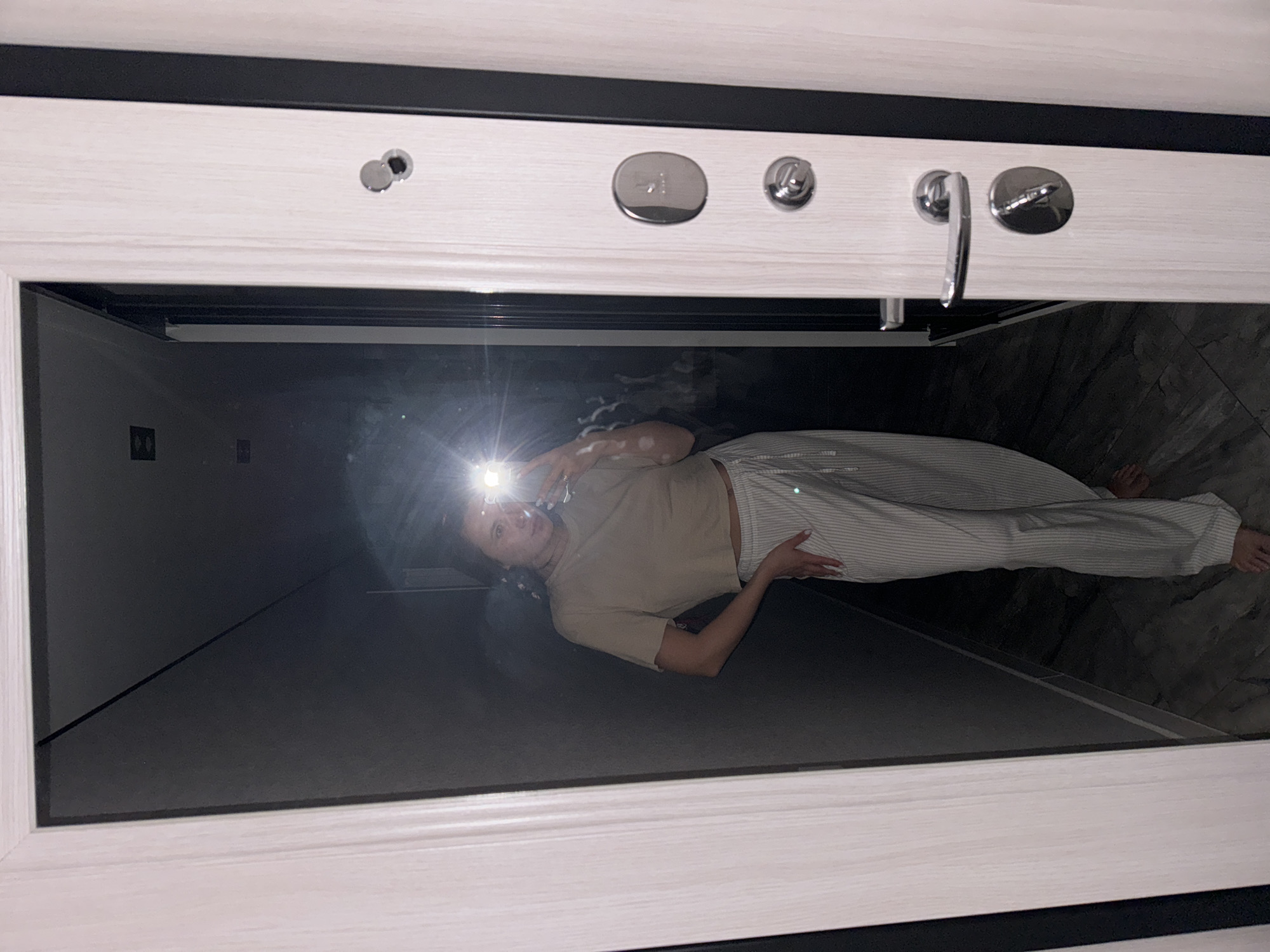 A woman is taking a selfie in a mirror. She is wearing a tan shirt and striped pants. The mirror is surrounded by a white frame with a black top. The location is Unknown, Unknown.