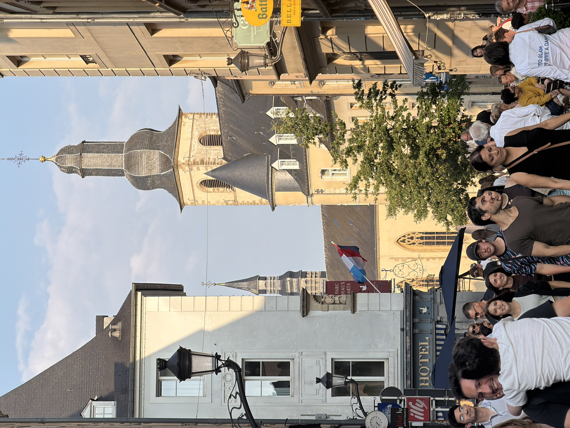 The image depicts a street scene in Luxembourg City, Luxembourg, featuring a blend of historic architecture and contemporary life. In the background, a prominent church tower with a dark, conical roof and a cross at its apex dominates the skyline, flanked by other traditional European buildings. The street is bustling with people walking and interacting, suggesting a lively urban atmosphere.
Several shops and establishments are visible along the street. A sign for "HOTEL" with an awning above indicates accommodation, while other signs for "illy" and a clock suggest commercial activity. In the foreground, people are walking in various directions, some engaged in conversation, others seemingly absorbed in their own thoughts. A man in a dark shirt is holding a child, who is leaning in to hug him, indicating a moment of affection.
The lighting suggests it is either late afternoon or early evening, with the sun casting long shadows and illuminating the buildings with a warm glow. A flag with red, white, and blue stripes is visible, possibly a nod to French influence or heritage. Trees are interspersed throughout the scene, adding a touch of nature to the urban landscape. The overall impression is one of a vibrant, historic European city during a pleasant time of day.