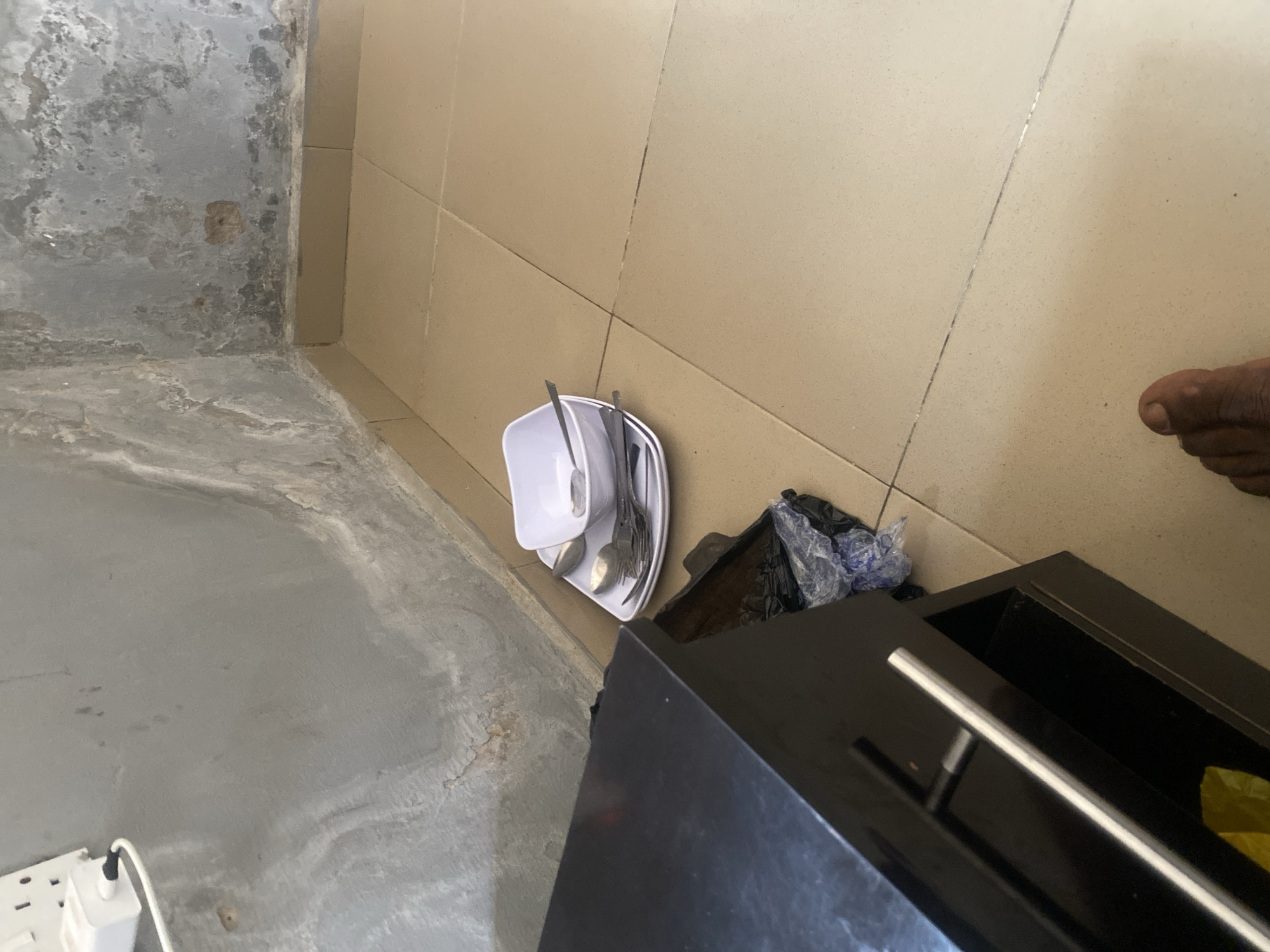 The image depicts a corner of a room, likely in Jalingo, Nigeria. The walls show signs of wear and potential dampness, with peeling paint and a textured, uneven surface. A beige tiled floor is visible, with grout lines forming a grid pattern. On the floor, near the corner and a dark wooden cabinet with a silver handle, is a stack of white plates, a white bowl, and several pieces of cutlery, including spoons and forks. A black plastic bag, possibly containing trash, is partially visible next to the cabinet. In the upper left of the image, a white electrical outlet with a plug and white cable is mounted on the wall. The lower right of the frame shows a portion of a person's bare foot. The lighting suggests it is daytime, possibly indoors with natural light.