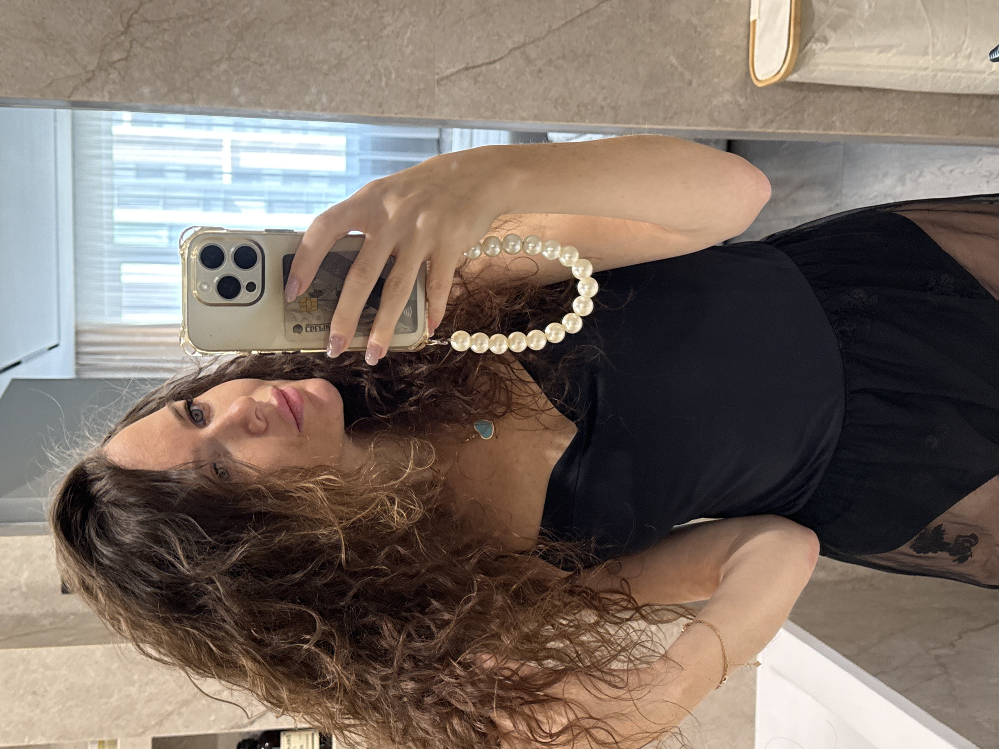 A woman with curly brown hair is taking a selfie in a bathroom with a marble counter and light-colored walls. She is holding a phone with a clear case adorned with a pearl wrist strap. The phone is covering part of her face, but her blue eyes are visible. She is wearing a black top and a necklace with a blue pendant. A window with blinds can be seen reflected in the mirror. The image does not provide information about the city of Khimki, Russia.