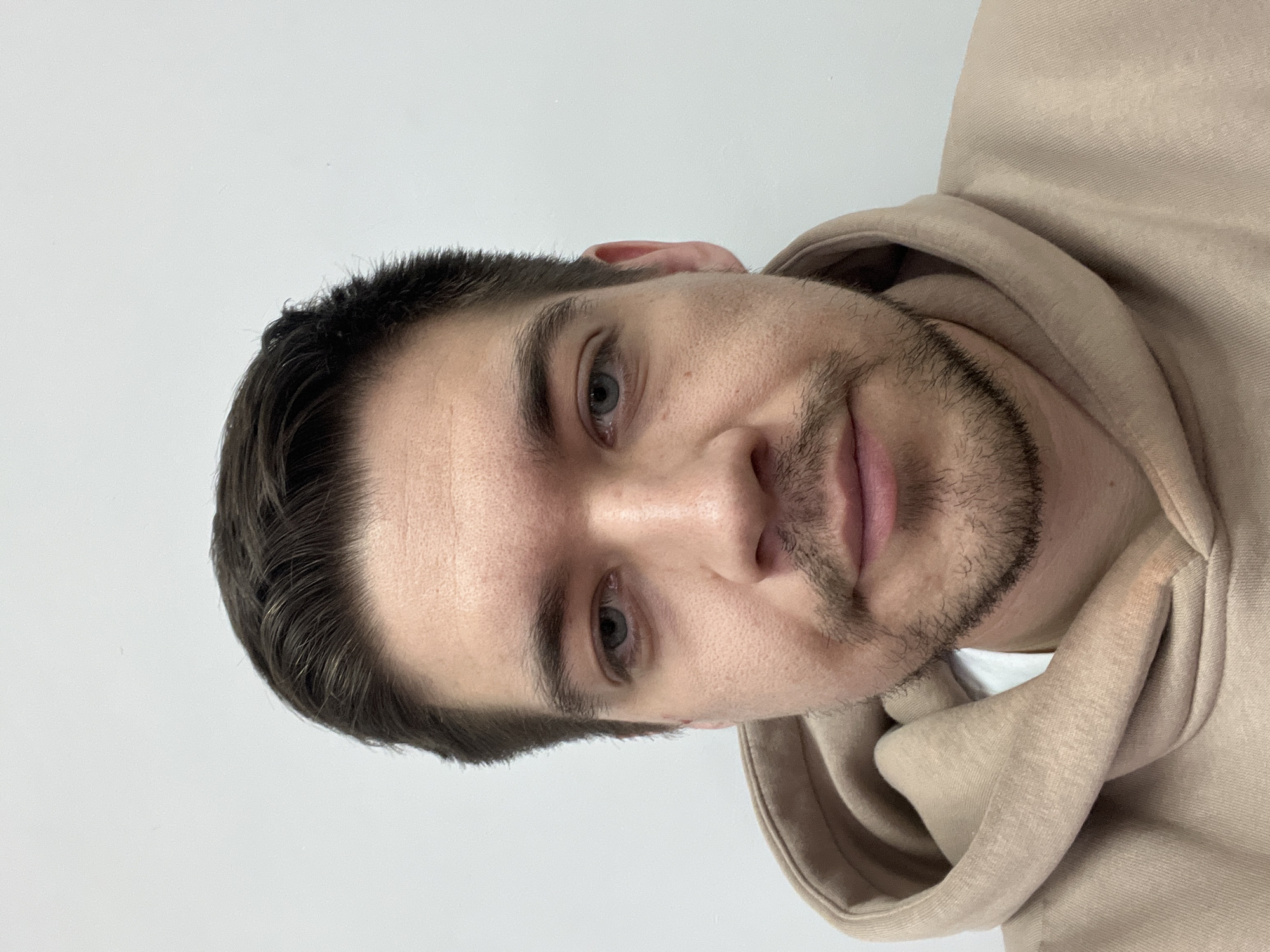 A close-up portrait features an adult man positioned against a plain, bright white background. The man's head and upper torso are visible, with his head appearing tilted due to the image orientation. He is looking directly forward, making eye contact with the viewer. He has short, dark brown hair, light-colored eyes (appearing blue or grey), and a short beard and mustache. His expression is neutral to slightly serious, with his lips slightly parted. He is wearing a light beige or tan-colored hoodie. The fabric of the hoodie is visible around his neck and shoulders, with folds and texture clearly discernible. The lighting is even and bright, suggesting an indoor setting. No other objects, people, or discernible text are present in the frame. This image was taken in Kazan, Russia.