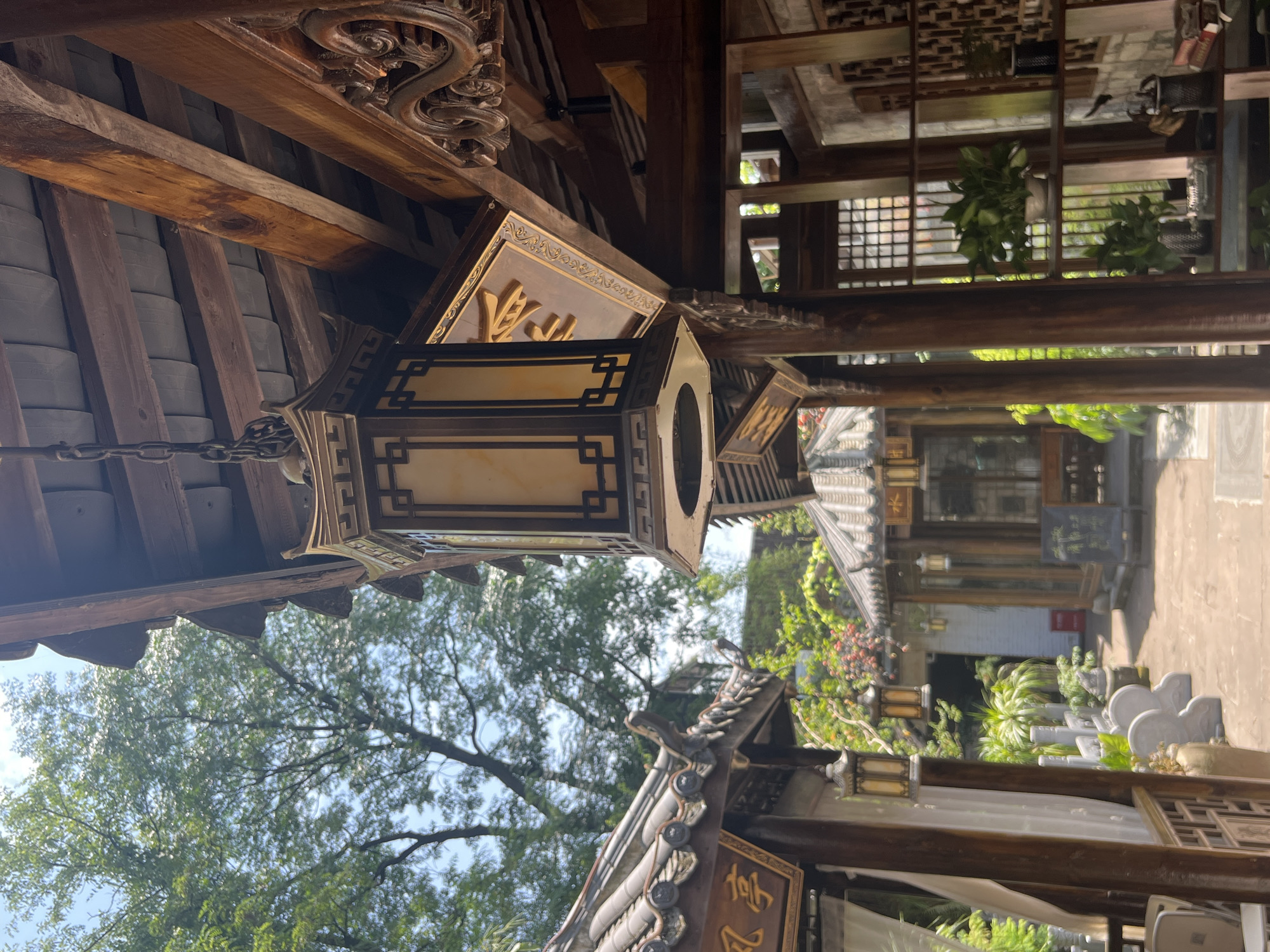 The image shows an outdoor scene with traditional Chinese architecture. In the foreground, a decorative lantern hangs from a wooden beam, suspended by a chain. The lantern is octagonal with ornate detailing and has a light-colored panel. To the left, a section of a building with a tiled roof and a wooden sign with Chinese characters is visible. Further back, a courtyard is visible with more traditional buildings, including a hexagonal pavilion with a tiled roof. Green trees and lush foliage surround the area. The ground appears to be paved. The lighting suggests a sunny day. The scene evokes a sense of tranquility and cultural heritage. The location is identified as Yuzhong District, China.