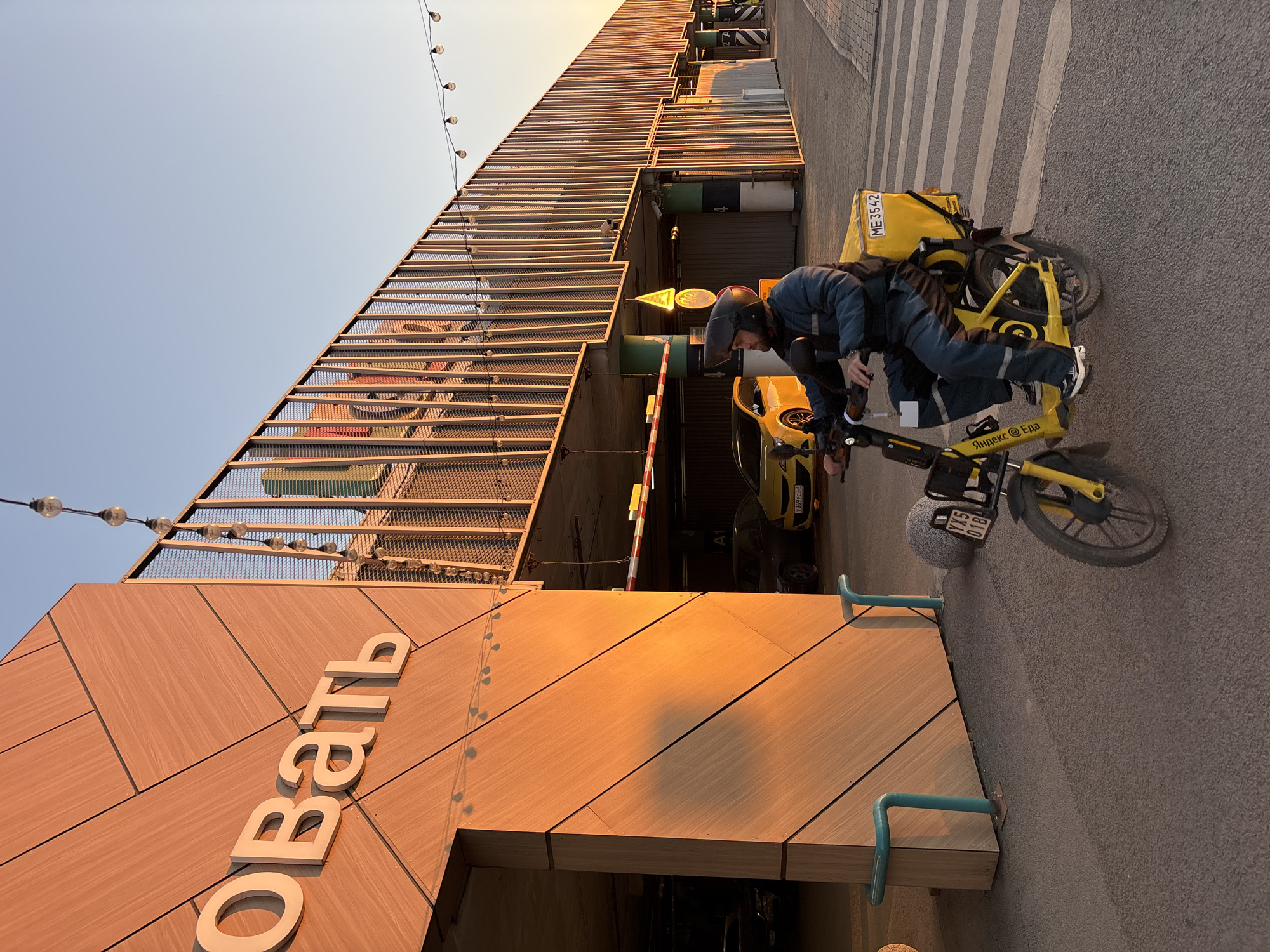 A delivery man on a yellow scooter with a "Yandex Eda" (Yandex Food) logo and "ME 3542" license plate number, is parked outside a building with "ОВАЦИЯ" (Ovation) written in large white Cyrillic letters on the exterior. The building has a facade of reddish-brown horizontally laid wood panels, with strings of lights above. The scooter and the delivery man, wearing a helmet and a blue jacket, are positioned on a grey pavement. Behind them is a parking garage entrance with a yellow car visible inside. Overhead are the metal beams of a parking structure. The location is Kudrovo, Russia.