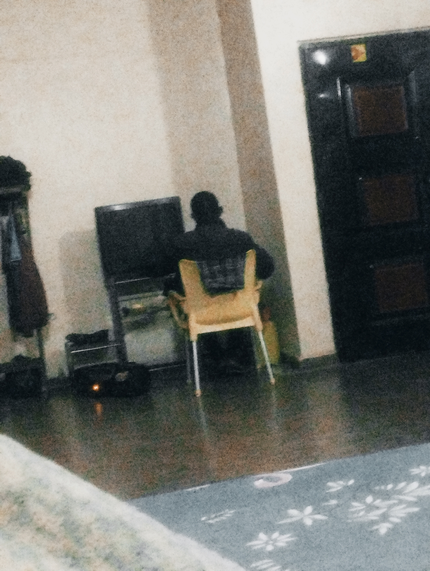 The image depicts an indoor scene, likely a room. In the center, a person is seated on a light-colored chair, facing away from the camera. The person appears to be wearing dark clothing. To the left of the person is a television set mounted on a stand. There are some items on the stand and on the floor in front of it. To the right of the person is a dark-colored door. The walls are a neutral color, and the floor appears to be tiled or carpeted in a dark hue. There is also a hint of a patterned rug in the foreground. The overall lighting is dim. There is no discernible connection to the city of Jos, Nigeria, in the visual elements of the image.