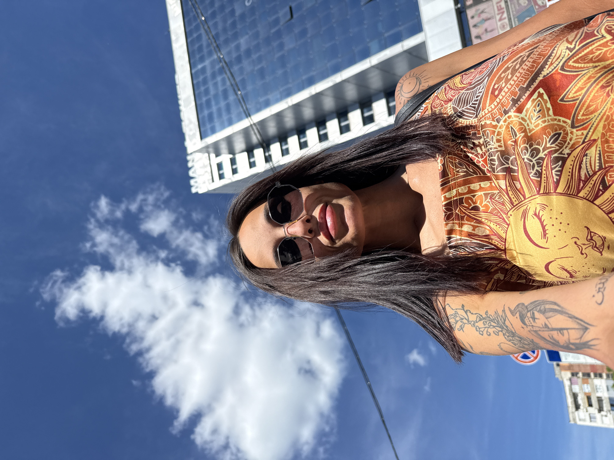 A woman is standing outside in Pleven, Bulgaria, beneath a partly cloudy sky and a modern glass-and-steel office building with a white frame. She has long, dark hair and is wearing sunglasses and a yellow-and-brown patterned top. She has multiple tattoos on her arms. A street sign is visible in the background.