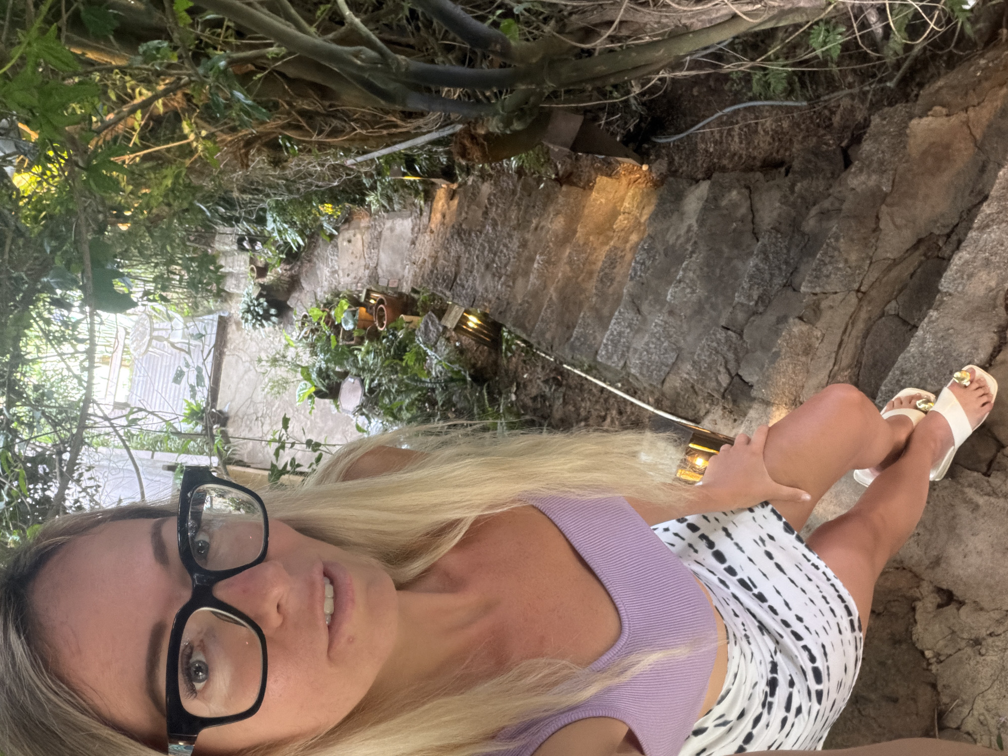 A woman with blonde hair and glasses is taking a selfie while descending a stone staircase in a lush, overgrown garden area. She is wearing a purple ribbed tank top and a black and white tie-dye skirt. The staircase is lined with potted plants and illuminated by small, warm lights. The location is identified as Xuân Thành, Vietnam.