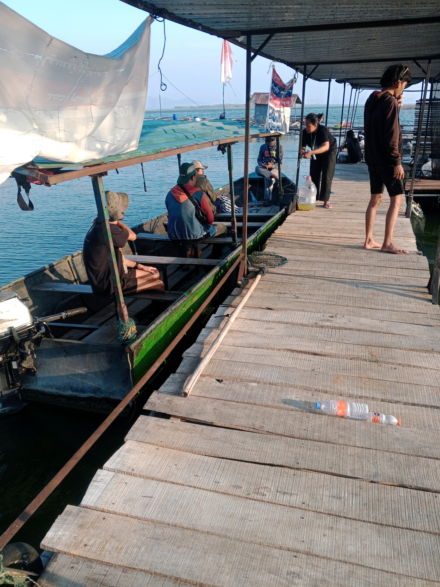 In Sendang, Indonesia, a wooden dock extends over a body of water, possibly a lake or river. Several people are present. On the dock, a man in a dark hoodie and shorts stands facing away from the camera, holding a cigarette. Nearby, a woman in a black t-shirt and pants stands next to a large jug of yellow liquid. A boat is docked alongside the wooden structure. Inside the boat, several individuals are seated. One person wears a hat and a black t-shirt, another a blue jacket over an orange shirt, and a third a light-colored hat and jacket. The boat has an outboard motor attached to its stern. The scene is illuminated by the warm light of what appears to be late afternoon or early morning sun, casting shadows on the wooden planks of the dock. Two plastic water bottles are visible on the dock, one a clear plastic bottle with an orange cap and the other partially visible with a white cap. A white tarp is draped over a structure near the boat.