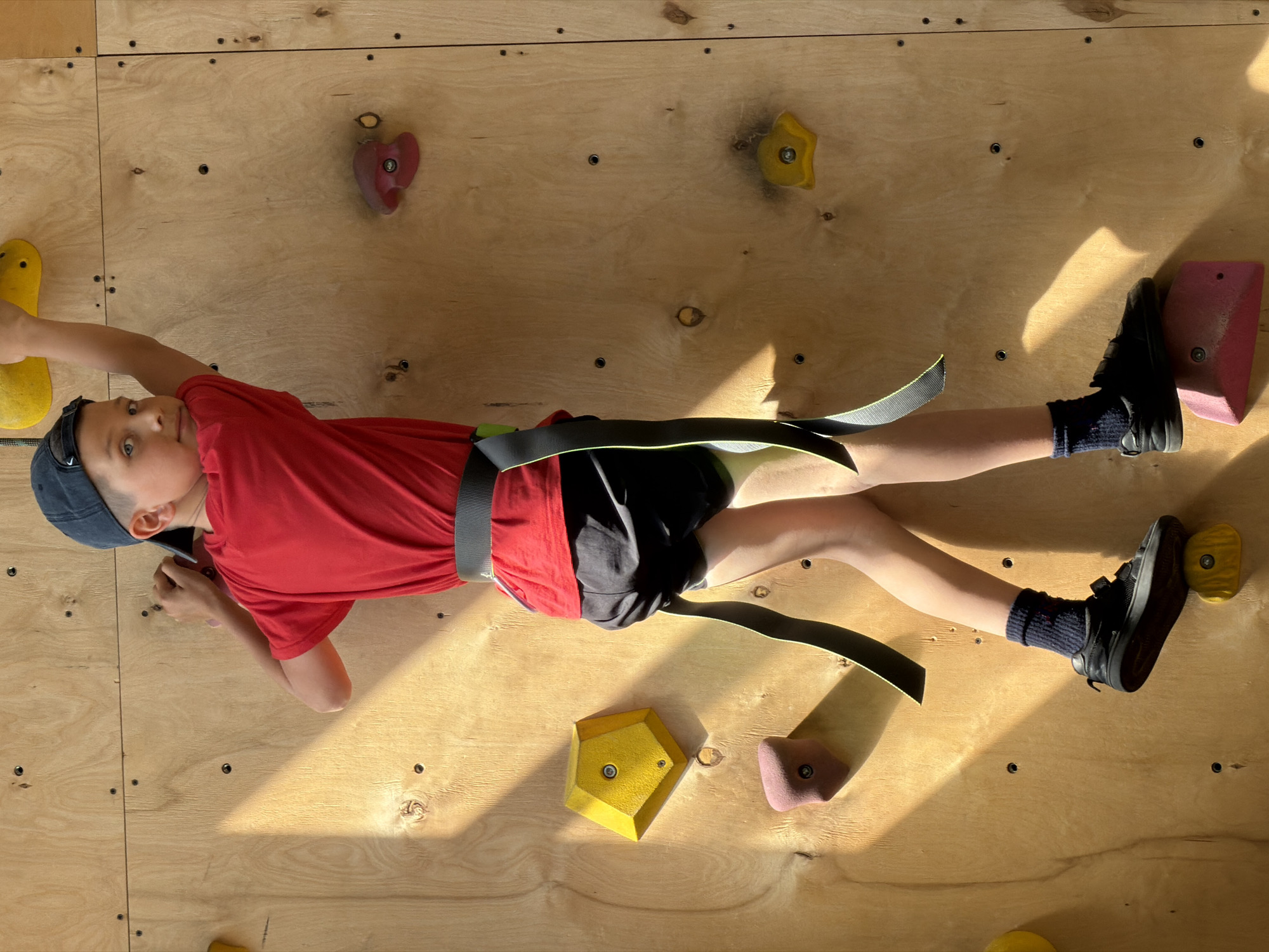 A child wearing a red shirt, black shorts, black shoes and a blue cap is lying on a climbing wall made of plywood with various colorful climbing holds attached. The child is wearing a climbing harness and is positioned at an angle, with their head towards the upper left corner of the frame. The wall appears to be indoors, illuminated by natural light creating shadows. The location is not possible to determine from the image, and the image does not contain any information that could identify Izhevsk, Russia as the location.