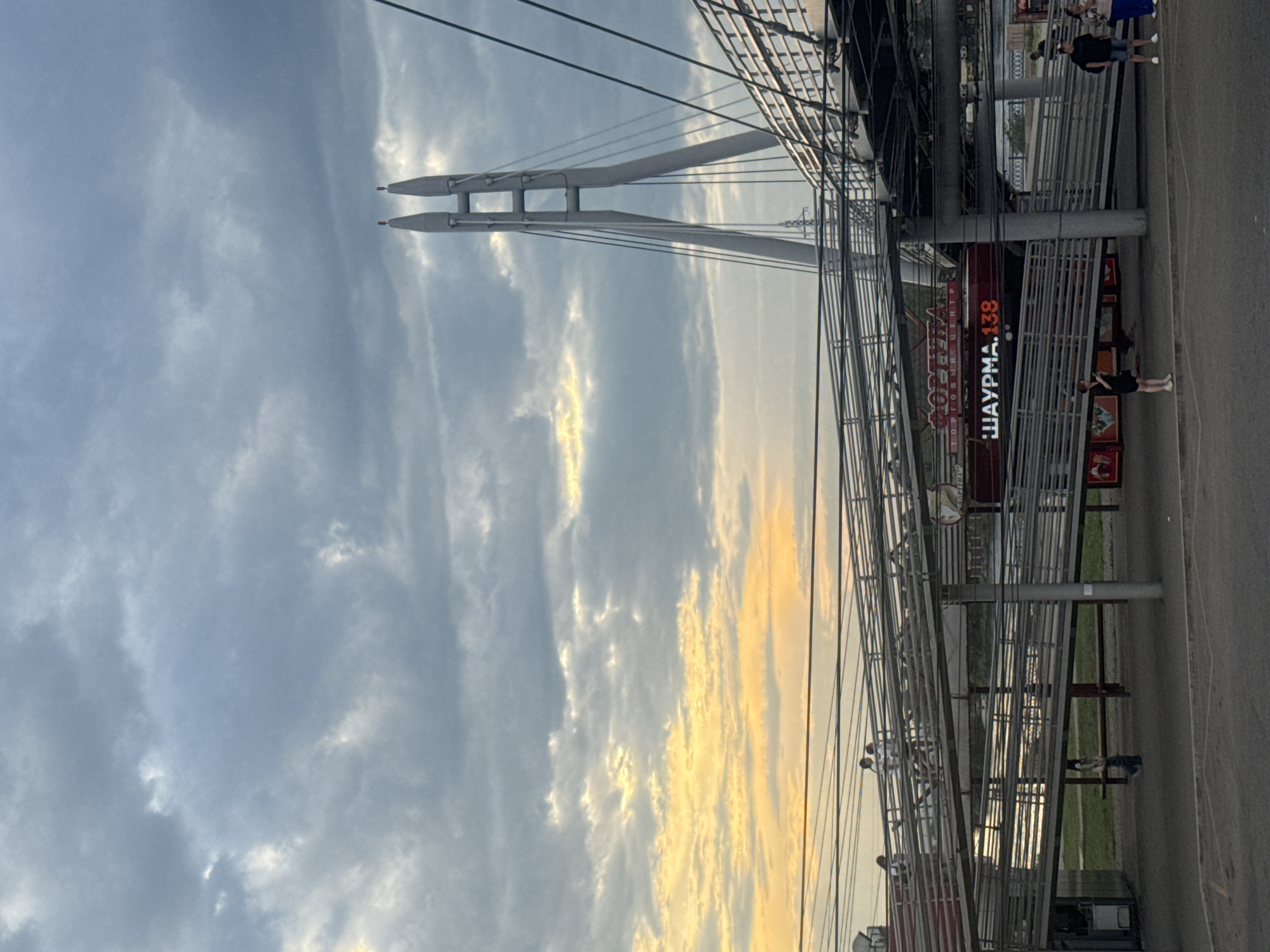 The image captures a pedestrian bridge in Irkutsk, Russia, with a cable-stayed bridge visible in the background under a cloudy sky tinged with sunset colors. The foreground features the ramp of the pedestrian bridge, with several individuals walking on it and at its base. A red building with illuminated signage, including the text "ШАУРМА. 138" and "ТОРГОВЫЙ ЦЕНТР," is situated beneath the bridge. The overall scene depicts an urban environment with elements of both infrastructure and commercial activity.