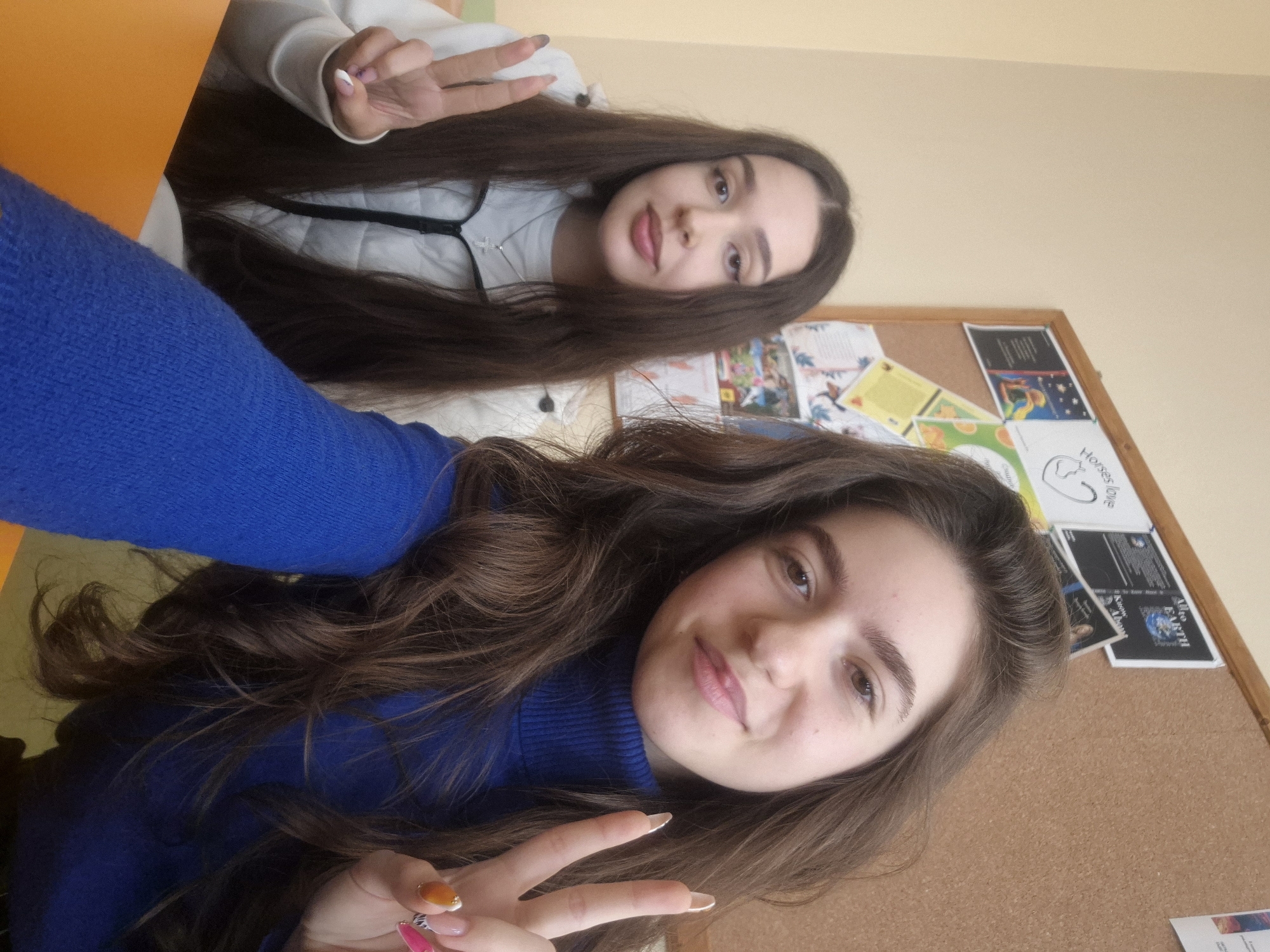 Two young women are posing for a selfie. The woman in the foreground is wearing a blue turtleneck sweater and displaying a peace sign. The woman in the background is wearing a white sweater and also displaying a peace sign. A bulletin board is visible in the background with various papers and images pinned to it. The photo was taken in a classroom, likely at a school in Pavlikeni, Bulgaria. An orange table edge is visible to the left.