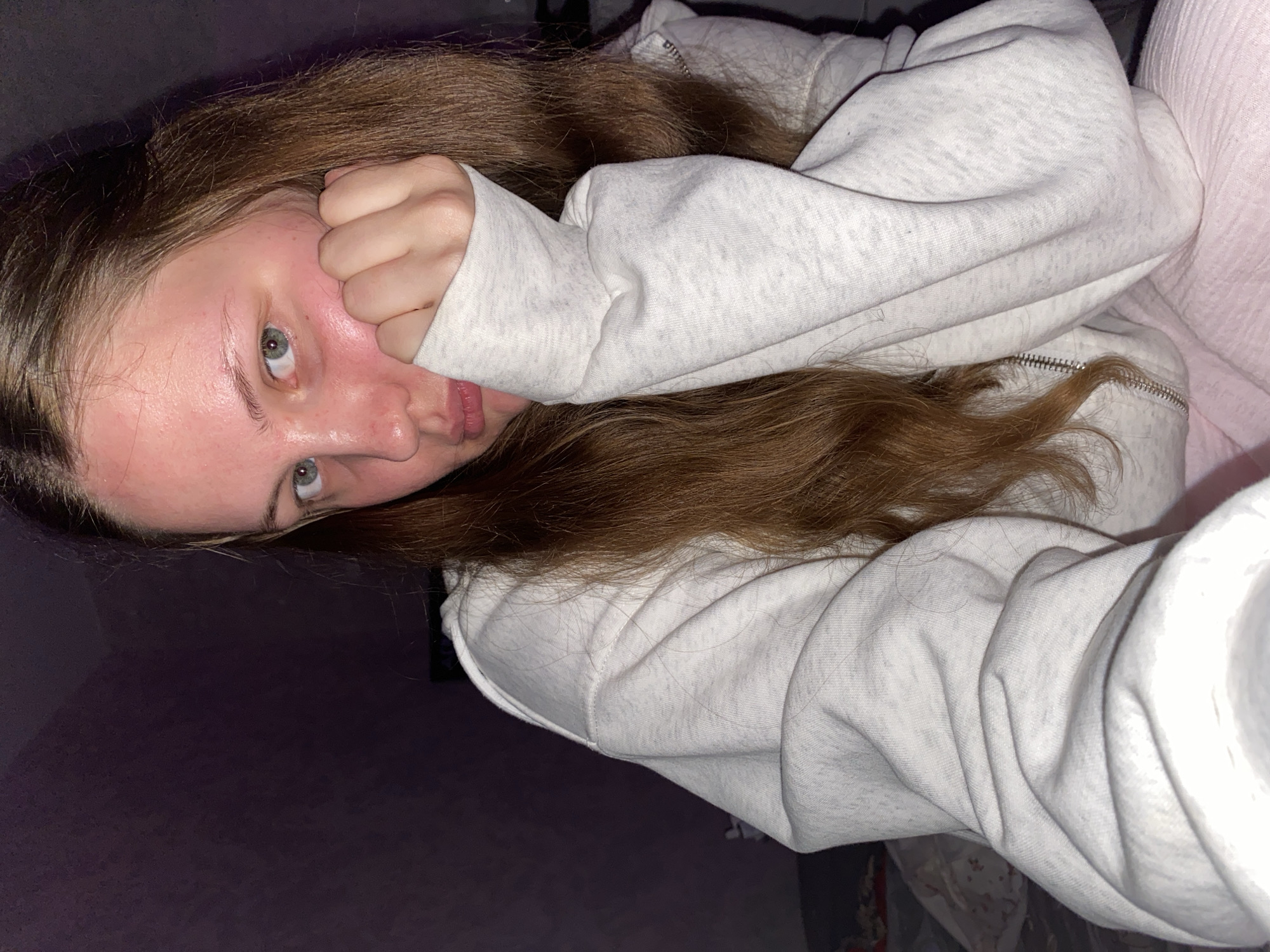 The image shows a young person with long brown hair and green eyes, resting their head on their hand. They are wearing a light grey zip-up hoodie and a pink garment underneath, possibly pajamas. The background is dimly lit with a purple hue, suggesting an indoor setting, possibly a bedroom. There is no discernible indication of the specific location within the city of Izhevsk, Russia, or any details that would suggest this context. The lighting is low, creating a soft and somewhat intimate atmosphere. The subject's expression is neutral to slightly melancholic.