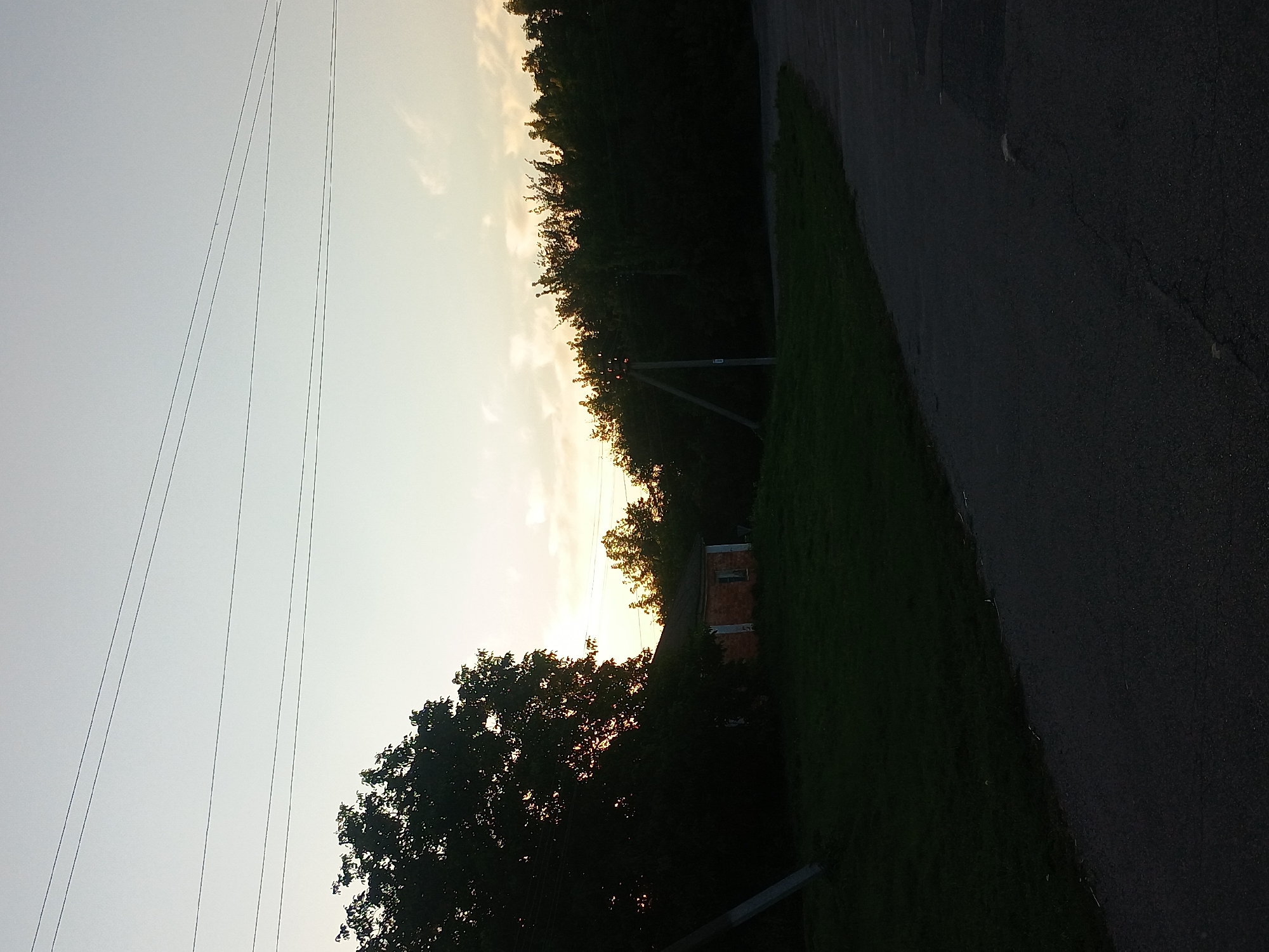 The image depicts an outdoor scene at dusk, likely in the village of Kliushnykivka, Ukraine, given its context. A dark asphalt road curves from the lower right of the frame towards the left. Adjacent to the road, on the left side of the image, is a grassy area. Partially visible within this grassy area and silhouetted against the fading light is a small, brick building with a dark roof and a single visible window. To the left of the building and extending further left is a large, dense tree, its dark foliage obscuring much of the background. In the distance, a line of dark trees forms a treeline across the horizon. Above the treeline, the sky transitions from a pale yellow-orange near the horizon to a light blue, with scattered wispy clouds. Several thin power lines are stretched across the upper portion of the sky. Two wooden utility poles are also present, one on the far left near the large tree, and another on the right, standing behind the treeline. The overall impression is one of a rural landscape at the end of the day.