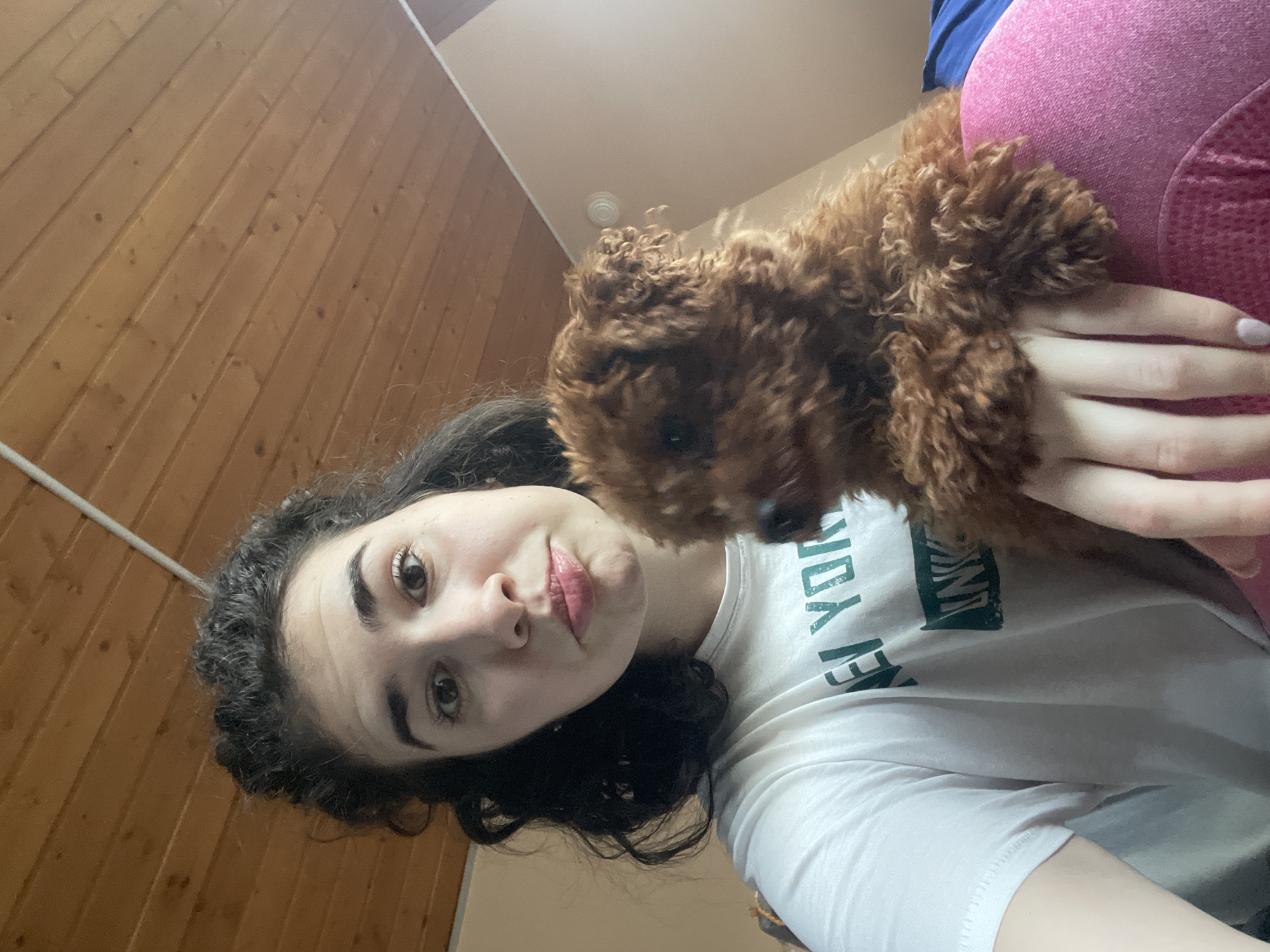 A young woman with dark curly hair and fair skin is holding a small, brown, curly-haired dog. She is wearing a white t-shirt with green text and has pink leggings behind her. The background includes wooden paneling and a white ceiling. The specific location within Sofia, Bulgaria, cannot be determined from the image.