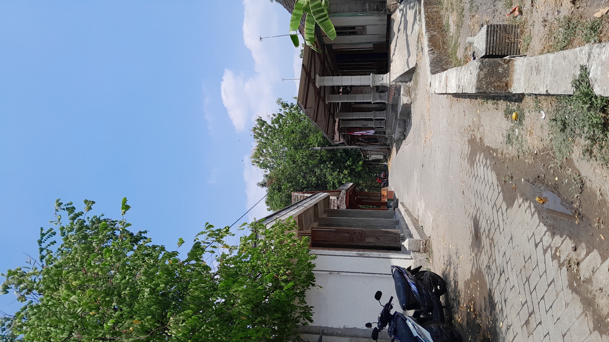 The image depicts a street scene in the city of Sumberarum, Indonesia, on a clear, sunny day. The perspective looks down a paved walkway that is lined with buildings and trees. On the left, a white building with a covered walkway is visible, with two motorcycles parked in front. Further down the street, more buildings with covered porches and pillars can be seen, some adorned with a red and white flag, possibly the Indonesian flag. Dense green trees provide a backdrop to the structures. The ground is a mix of paved brick and dirt, with some puddles and scattered debris. The sky is a bright blue with a few wispy clouds. The overall impression is of a quiet, residential street in a tropical climate.