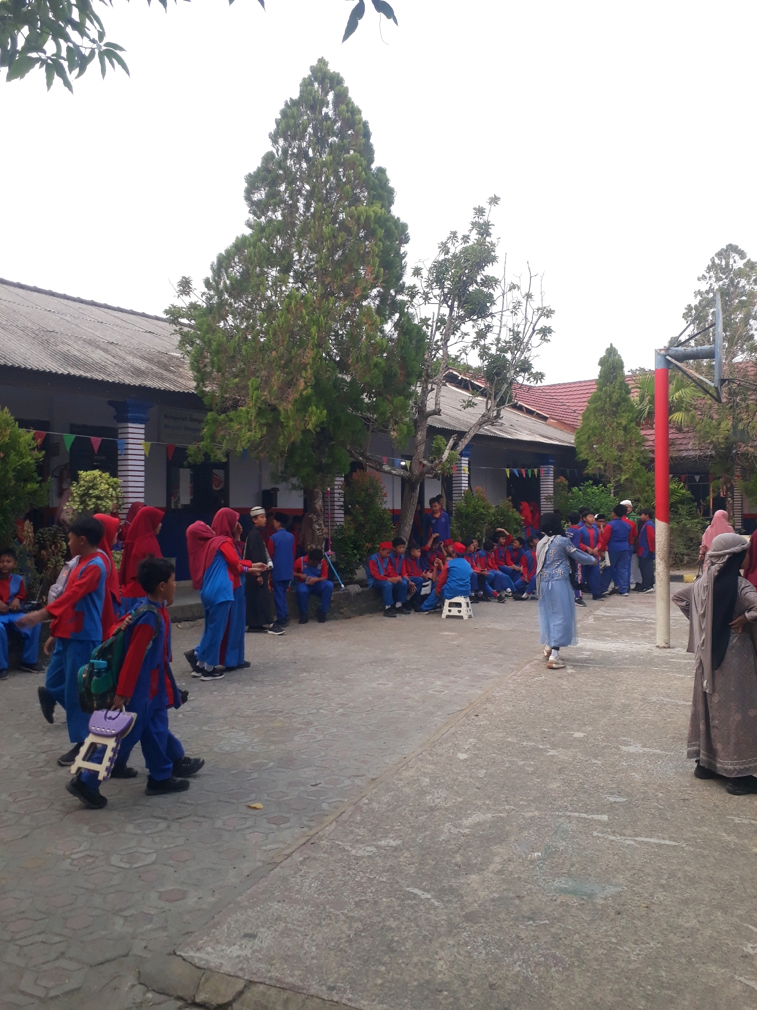 The image depicts a scene outside a school building, likely in the city of Pangkalpinang, Indonesia. A group of children, predominantly in red and blue school uniforms, are gathered in an outdoor courtyard. Some children are standing and conversing, while others are sitting on the ground or on small plastic stools. One child is carrying a small purple stool. Several adults are also present, some of whom are wearing headscarves. A basketball hoop is visible in the background. The school building has a corrugated metal roof and appears to be a single-story structure. Trees and foliage are present around the building and in the courtyard. The ground is paved with hexagonal and square concrete tiles. The overall atmosphere appears to be casual and possibly related to a school event or break time.