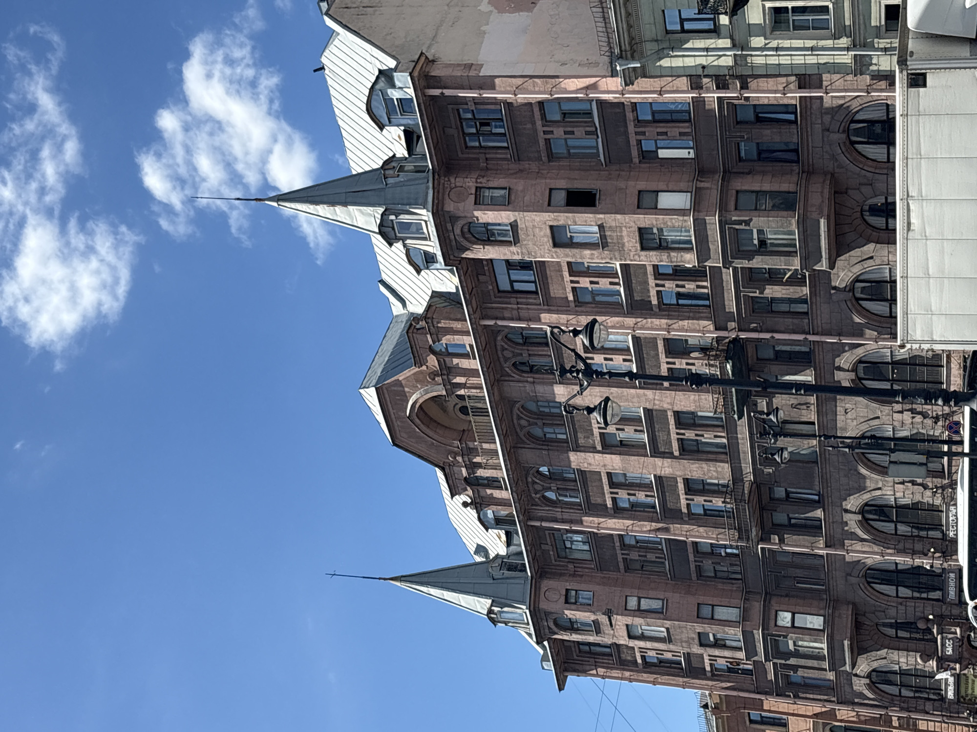 The media file is a vertical, low-angle, exterior shot of an ornate, multi-story building on a clear, sunny day. The building's facade is made of pinkish-brown stone and features numerous windows, balconies, and decorative architectural elements. Two pointed, grey spires rise from the roofline, adding to the building's imposing presence.
The sky is a vibrant blue with scattered white clouds, suggesting it is daytime. The sunlight casts shadows, highlighting the building's intricate details. The composition emphasizes the height and grandeur of the architecture.
Visible text on signs on the building's lower floors includes "РЕСТОРАН" (Restaurant) and "ПИВОМ" (Beer), indicating commercial establishments. Another sign is partially visible with Cyrillic script. The overall impression is of a historic and elegant urban environment. No people are visible in the frame, and the scene appears to be a still moment, not capturing any specific activity. The time of day appears to be mid-morning to afternoon, given the bright sunlight and clear sky.