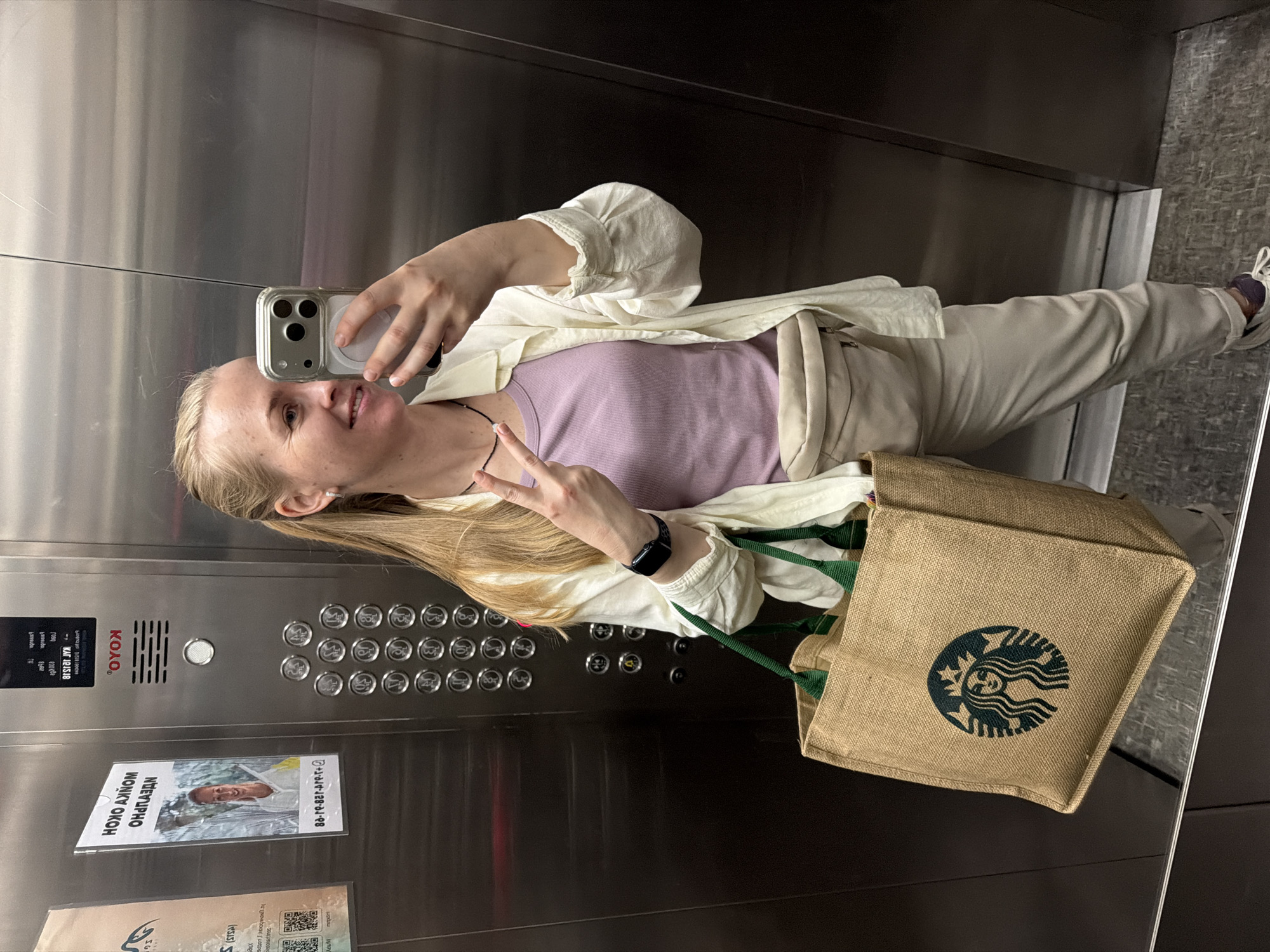 A blonde woman with a light-colored shirt and pants, and a fanny pack, stands in an elevator, taking a selfie with her phone. She holds a large brown tote bag with a Starbucks logo in her left hand and flashes a peace sign with her right hand. The elevator has stainless steel walls and a panel of buttons. A poster with Russian text and an image of a woman is visible on the left wall of the elevator. The floor of the elevator is a gray speckled pattern. The image appears to have been taken in the city of Khabarovsk, Russia, as suggested by the poster in the background which contains Cyrillic script, consistent with Russian signage.