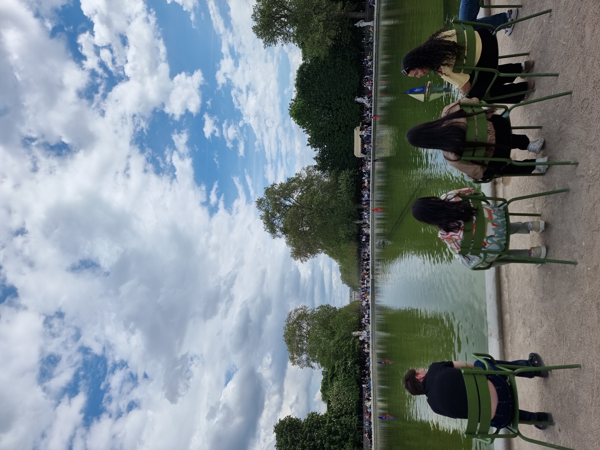 The image captures an outdoor leisure scene in what appears to be a public park, likely in Paris, France, given the distinctive green chairs. The perspective is from the side, with a large, rectangular pond dominating the middle ground, flanked by a gravel path and lush trees.
In the foreground, four individuals are seated on iconic green metal garden chairs, positioned along the edge of a gravel path that runs beside the pond. They are facing the water, observing the activity. One person with long, dark, curly hair, wearing a yellow top and dark pants, is seen intently looking towards a small model sailboat with a blue and white sail floating near the pond's edge. Another person, seen from behind, also has long dark hair and wears a light-colored jacket. Further down the row, two more individuals are seated, one in a patterned shirt and another in a black top, both seemingly engaged with the scene on the water.
The calm surface of the large pond reflects the trees and sky, appearing a greenish hue. Several model sailboats are visible on the water, indicating a popular recreational pastime. The opposite bank of the pond is lined with many people, suggesting a bustling and well-attended park. Beyond the crowd, a dense row of green trees stretches into the distance, with a hint of architectural structures visible through and above the foliage.
The sky above is bright and partly cloudy, with patches of clear blue interspersed with white, fluffy cumulus clouds, suggesting a pleasant daytime setting. The overall atmosphere is one of relaxation and enjoyment in a classic park environment. No discernible text is visible within the image.
