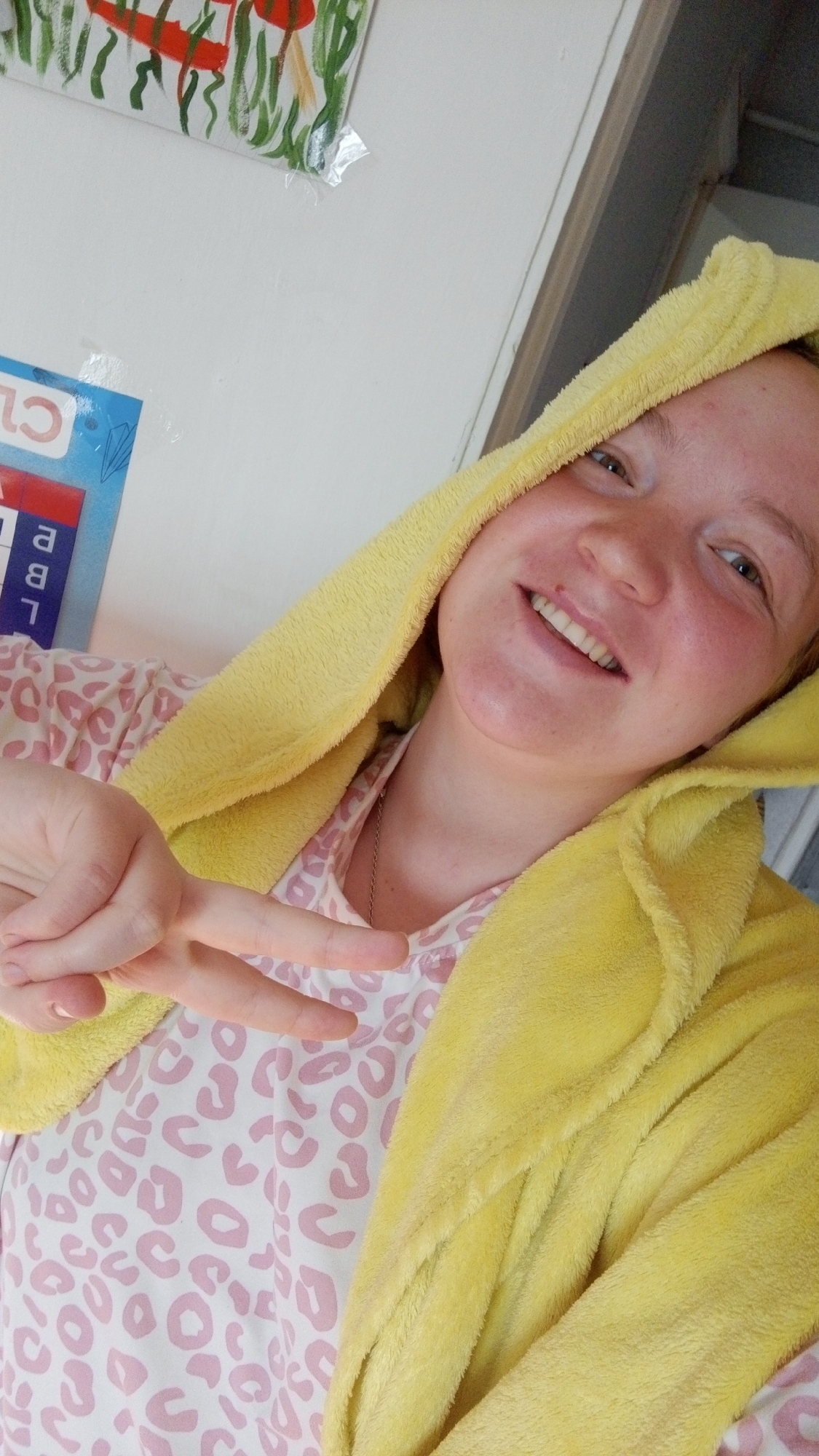 A woman wearing a yellow hooded robe and a pink and white patterned top is smiling and making a peace sign. The background includes a white wall with a small drawing and what appears to be a calendar or chart. The location is Unknown, Unknown.