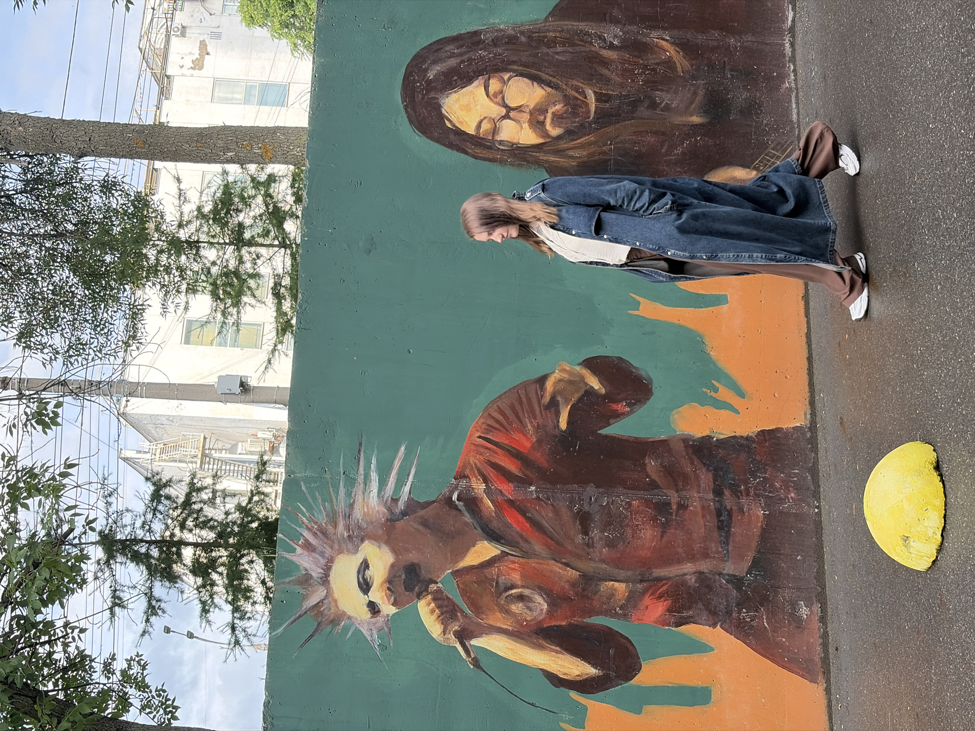 An outdoor shot shows a woman walking in front of a mural on a green wall. The mural features two stylized portraits: one of a man with spiky hair and a microphone, and another of a man with long hair and glasses. The woman, seen in profile from the right, wears a long denim coat over a white top and brown pants, with white sneakers. She is walking towards the right side of the frame. The wall is graffiti-covered and appears to be in an urban environment, with trees and buildings visible in the background. A yellow, dome-shaped object is on the wet asphalt in the foreground. This mural is located on an exterior wall of the "Chum" Youth Center in Izhevsk, Russia, at Komsomolsky Prospekt, 189.
