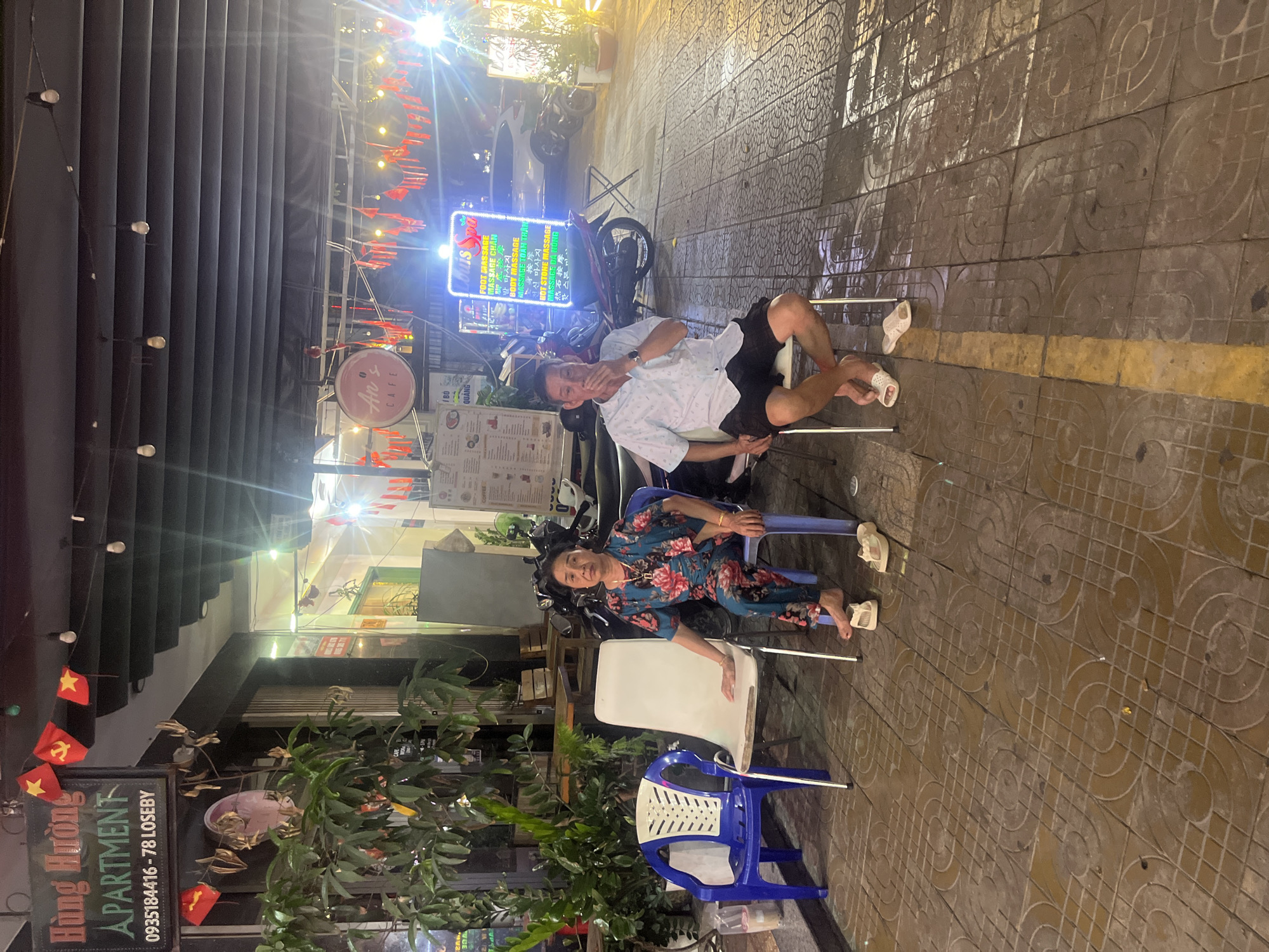 Two individuals are seated on chairs outside a business at night. The woman on the left, wearing a floral-patterned jumpsuit and sandals, is seated on a blue chair. To her right, a white chair is occupied by a white plastic bin. To the right of the white chair, a man in a light-colored patterned shirt and dark shorts, wearing sandals, is seated on a similar white chair. He is covering his mouth with his hand. Behind the individuals, a neon sign displaying "Louis Spa" with offerings like "FOOT MASSAGE" and "HOT STONE MASSAGE" is prominently visible. The ground is paved with patterned tiles, and there are potted plants to the left of the scene. A sign for "HUNG HUONG APARTMENT" with a phone number and address is visible above and to the left of the woman. The setting appears to be on a street in Da Nang, Vietnam, characterized by the architecture, signage, and the general atmosphere of an urban environment at night.