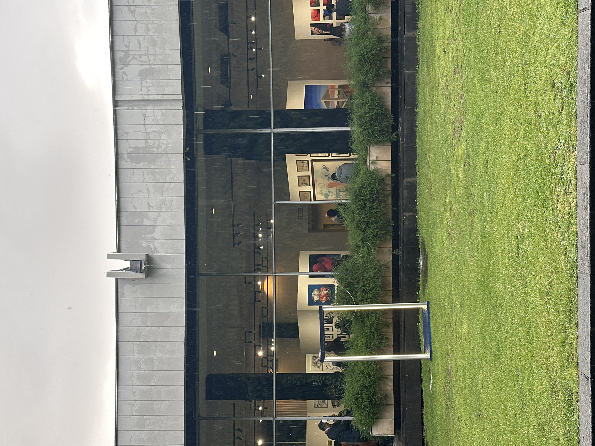 The photograph captures a section of the exterior of the Zurab Tsereteli Museum of Modern Art (MOMA Tbilisi), located in Tbilisi, Georgia. The modern building features a facade of gray tiled panels and large glass windows. The reflection in the windows reveals artwork displayed inside the museum, including paintings and sculptures. People are visible inside, observing the art. In front of the building, a well-maintained green lawn and a row of shrubs create a barrier between the building and the grass. A rectangular metal frame, potentially part of an outdoor installation or decorative element, stands on the lawn. The sky is overcast.