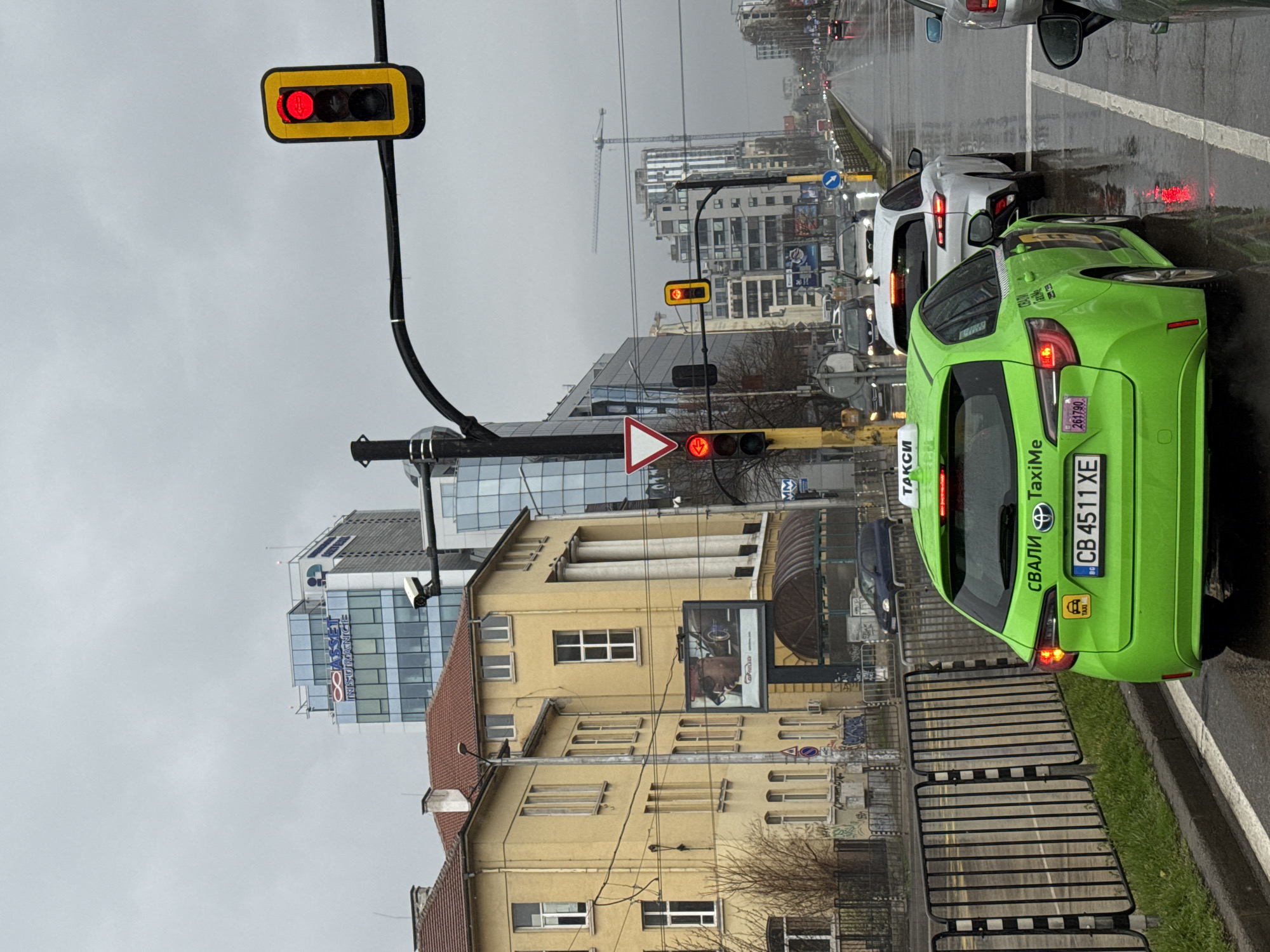 The image depicts a street scene in Sofia, Bulgaria, on an overcast day. It appears to be taken from inside a vehicle. In the foreground, a bright green taxi, identified by its color, taxi signage, and license plate "CB 4511 XE", is stopped at a traffic light. A white car is directly in front of the taxi. The traffic light in the immediate foreground shows a red light, indicating that traffic is stopped. Further down the road, another traffic light is visible, also showing red.
Buildings line the street. On the right, a light yellow building with multiple windows and a red tile roof is visible. A large billboard is attached to the building. Across the street, modern office buildings with glass facades can be seen, one of which displays the logo of "ASSET INSURANCE."
A construction crane is visible in the distance, suggesting ongoing construction in the area. The street is wet, indicating recent or current rain.
The general location is likely in a commercial or business district of Sofia, given the mix of older and modern buildings and the presence of business signage.