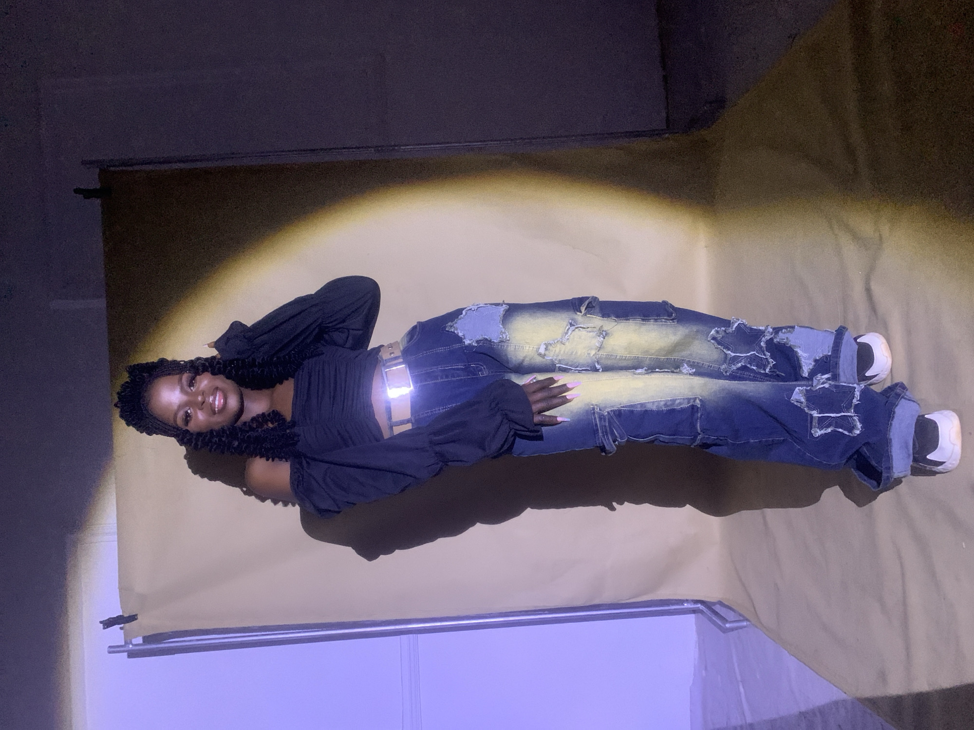 A woman lies on a light brown backdrop. She is wearing a black top with long, billowy sleeves, blue jeans with distressed and bleached details, a silver belt, and white shoes. Her hair is styled in long, curly braids. The backdrop is illuminated by a spotlight, casting a shadow of the woman to the left. The upper left and lower left corners of the image reveal parts of what appears to be a studio setup with dark walls and a support structure for the backdrop. The location is unknown.