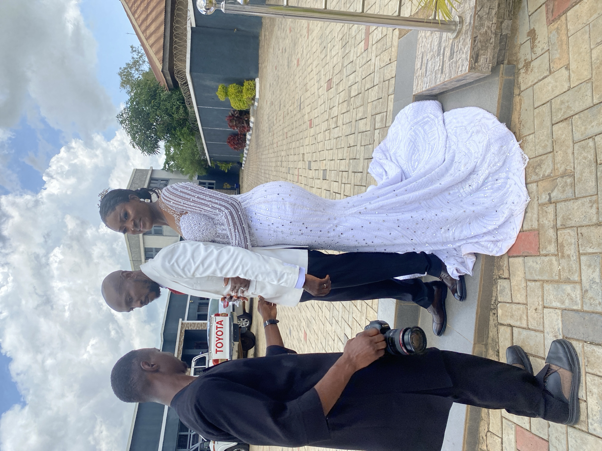 In Bukuru, Nigeria, a wedding photographer, dressed in a black traditional outfit, holds a camera and stands facing a wedding couple. The groom wears a white suit and the bride is in a white sequined mermaid wedding dress. They are positioned on a raised platform, and the bride is looking towards the camera. Behind them, a white Toyota pickup truck is visible, with buildings and trees in the background under a cloudy sky. The ground is paved with interlocking bricks.