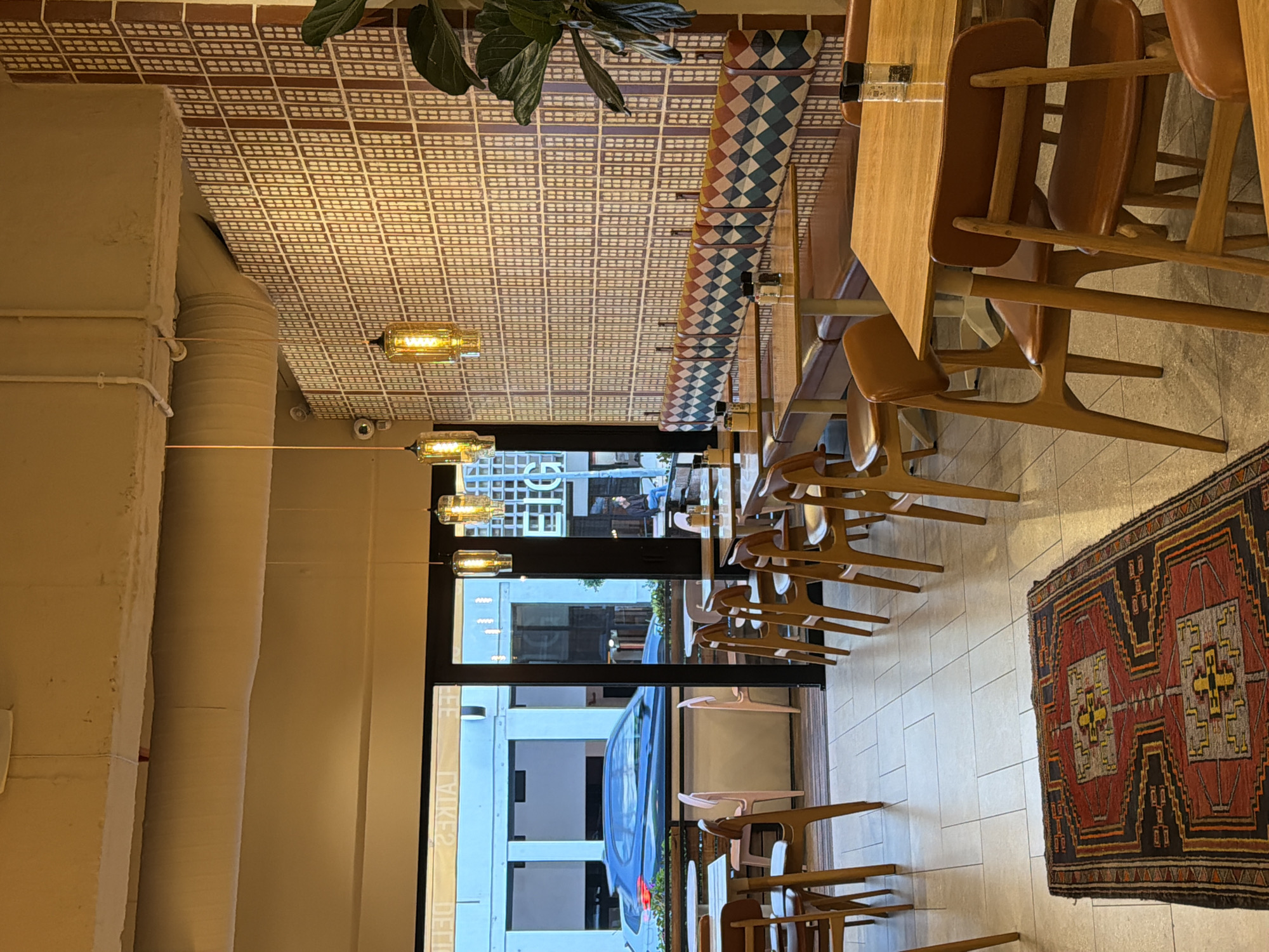 A view of a restaurant interior. The interior has long wooden tables and wooden chairs. A blue car is visible parked outside. The ceiling has geometric patterns. Lights hang from the ceiling. A rug with geometric patterns is on the floor.
The image shows details that enable its location in Cape Town, South Africa. The view through the window showcases architectural features and building styles typical of the city. Additionally, parked vehicles outside the restaurant offer insights into local transportation. "DELIKE2" on the window is a reference to "LIKE2 DELI," a restaurant located in Cape Town.