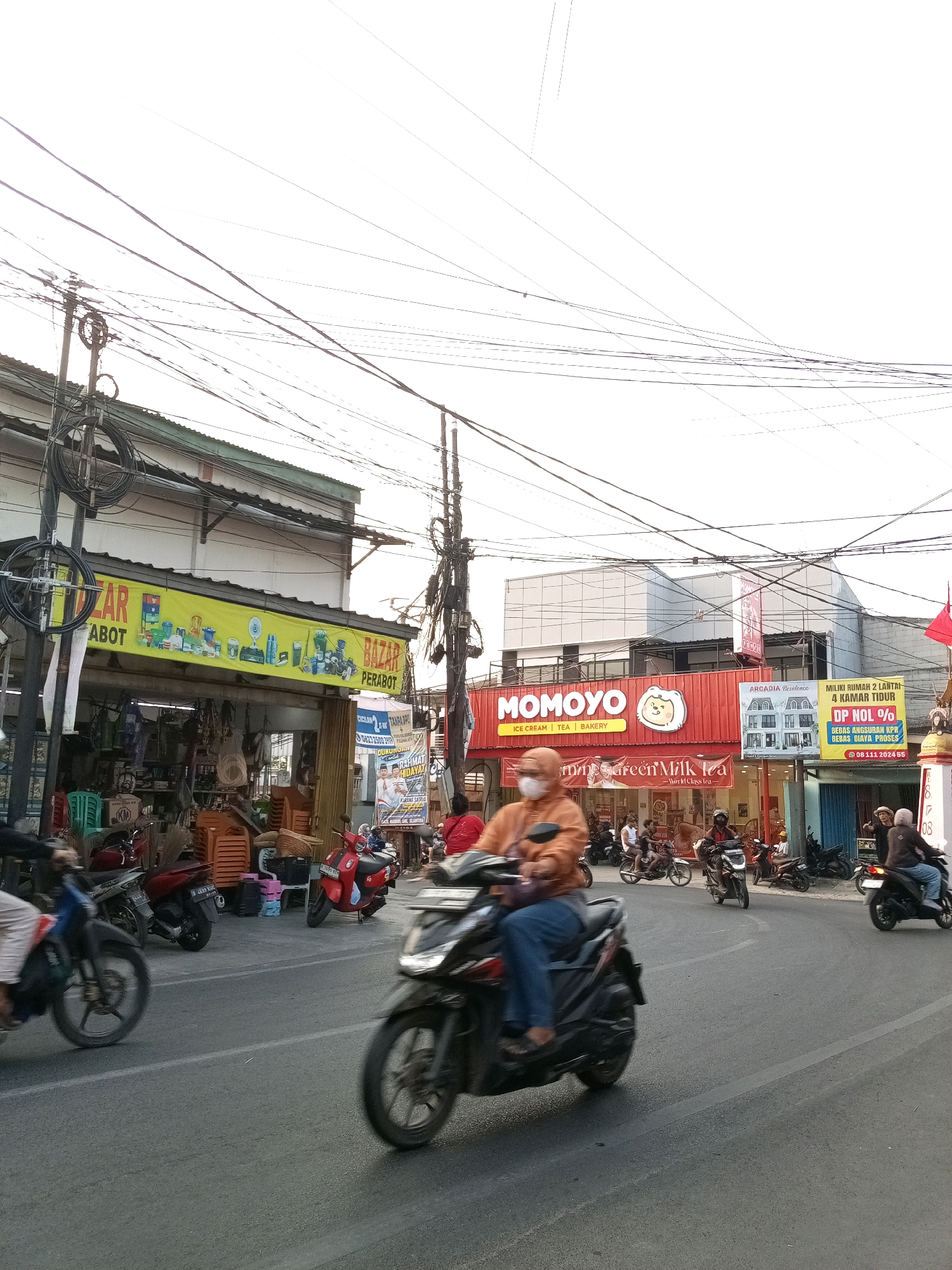 This image depicts a street scene in Karangsatria, Indonesia, with a focus on storefronts and traffic. In the foreground, several motorcycles are in motion. One motorcycle on the left is ridden by a person wearing an orange jacket and blue pants, with their face covered by a mask. Another motorcycle in the center is ridden by a person in a dark helmet. To the right, a motorcycle with two passengers is visible.
The background features a row of shops. On the left is a shop with a yellow awning that reads "BAZAR PERABOT," displaying various household items like appliances and cleaning supplies. Next to it is a shop with a red facade that says "MOMOYO" with the tagline "ICE CREAM | TEA | BAKERY" and a cartoon illustration of a bear. A smaller advertisement for "Arcadia Residence" is also visible. Further down the street, another shop has a red banner and a sign that appears to be a lottery or promotion, with numbers "08111 202455" prominently displayed.
The environment is characterized by a bright, overcast sky, numerous power lines crisscrossing overhead, and streetlights. The road surface is asphalt with painted lane dividers. The overall impression is a busy urban street during daylight hours.