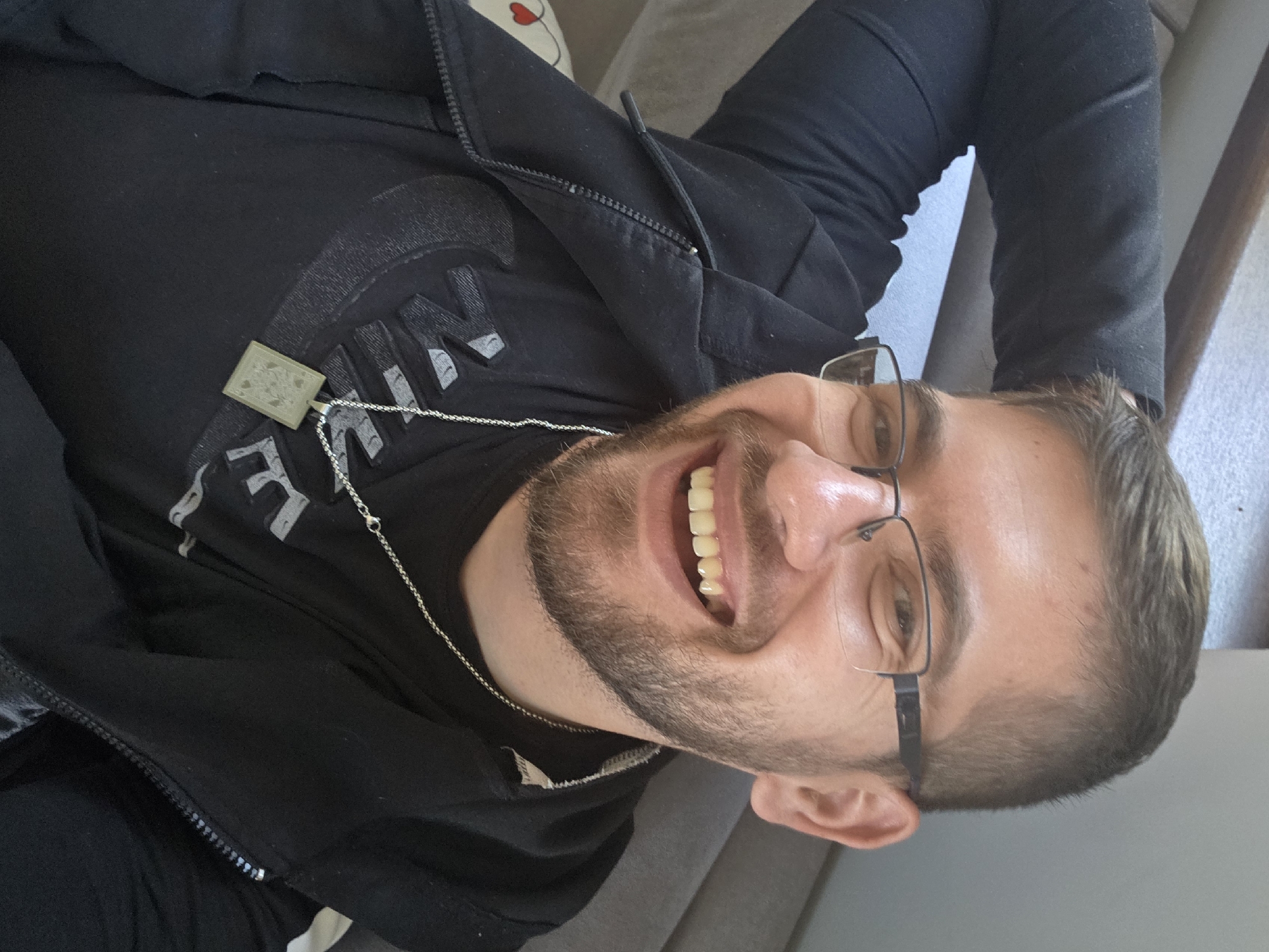 A man lies on a grey couch. He wears black framed glasses, a black zip-up jacket and a black Nike shirt with a silver pendant necklace. He is smiling broadly. The location is unspecified; there is no indication of Yagodovo, Bulgaria.